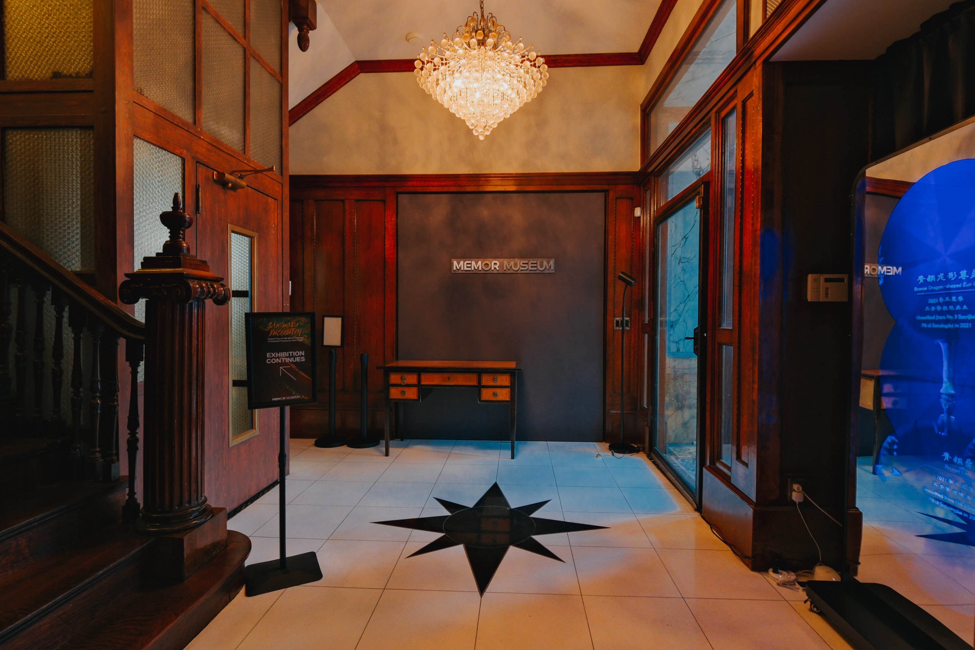 The entrance of a museum features a chandelier, wooden paneling, and a large star design on the floor.