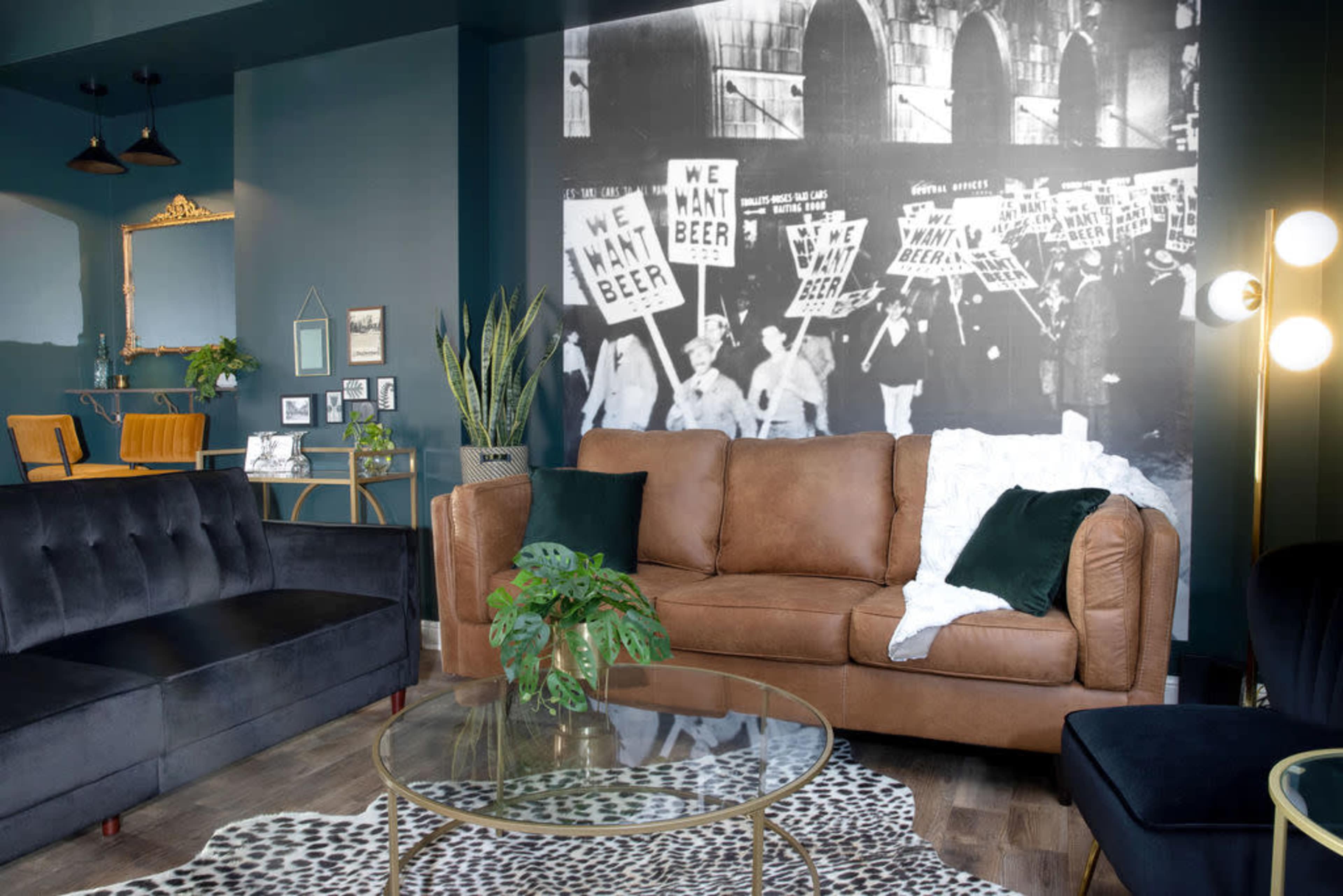 A stylish living room with a brown leather sofa, glass coffee table, and a large black-and-white wall mural depicting a protest for beer.