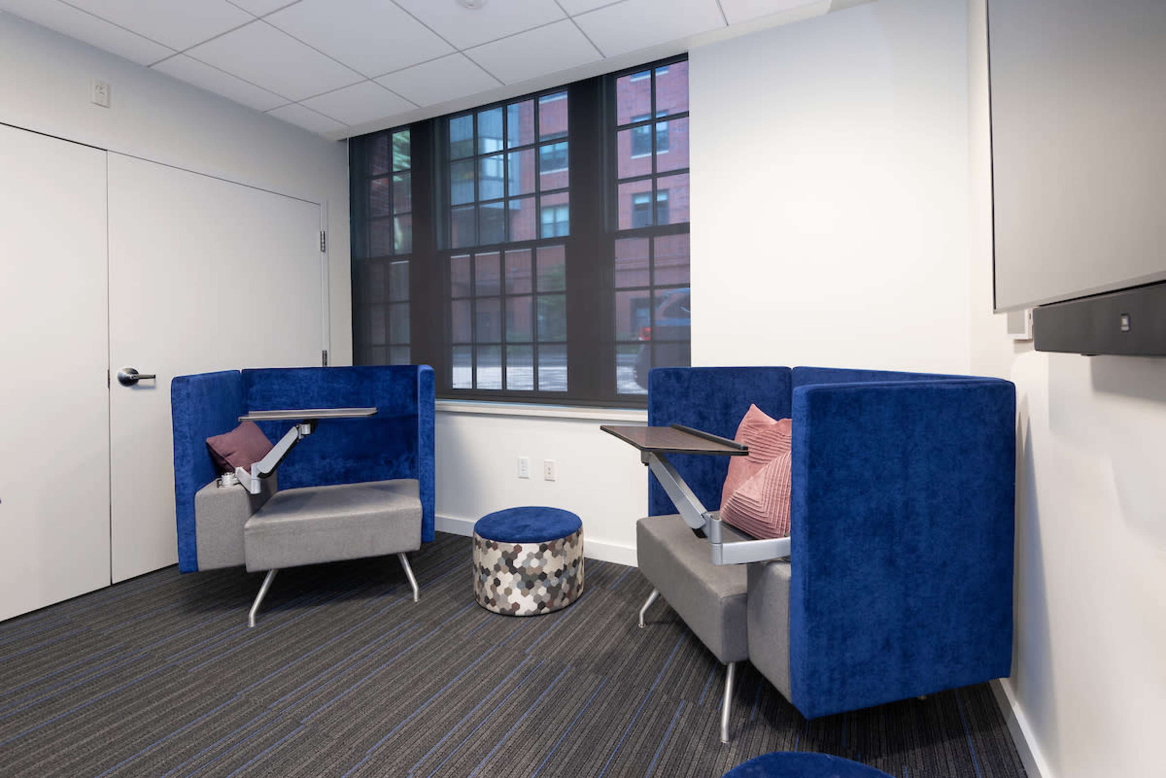 A corner of an office space featuring two blue upholstered seating areas with small folding tables and a round, patterned ottoman.