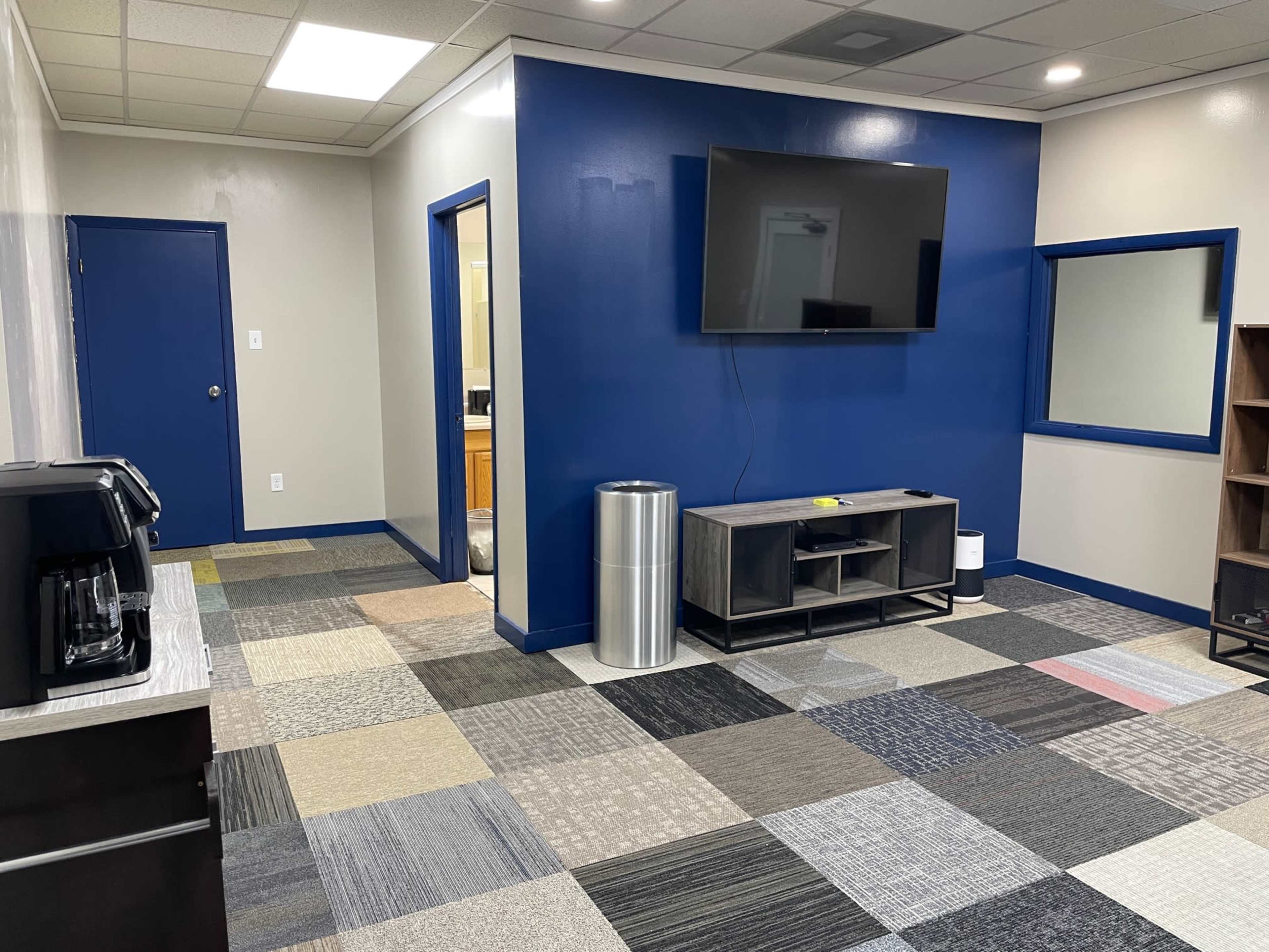 The room features a modern design with a television mounted on a blue wall, a coffee machine, and a combination of carpet tiles in various colors.