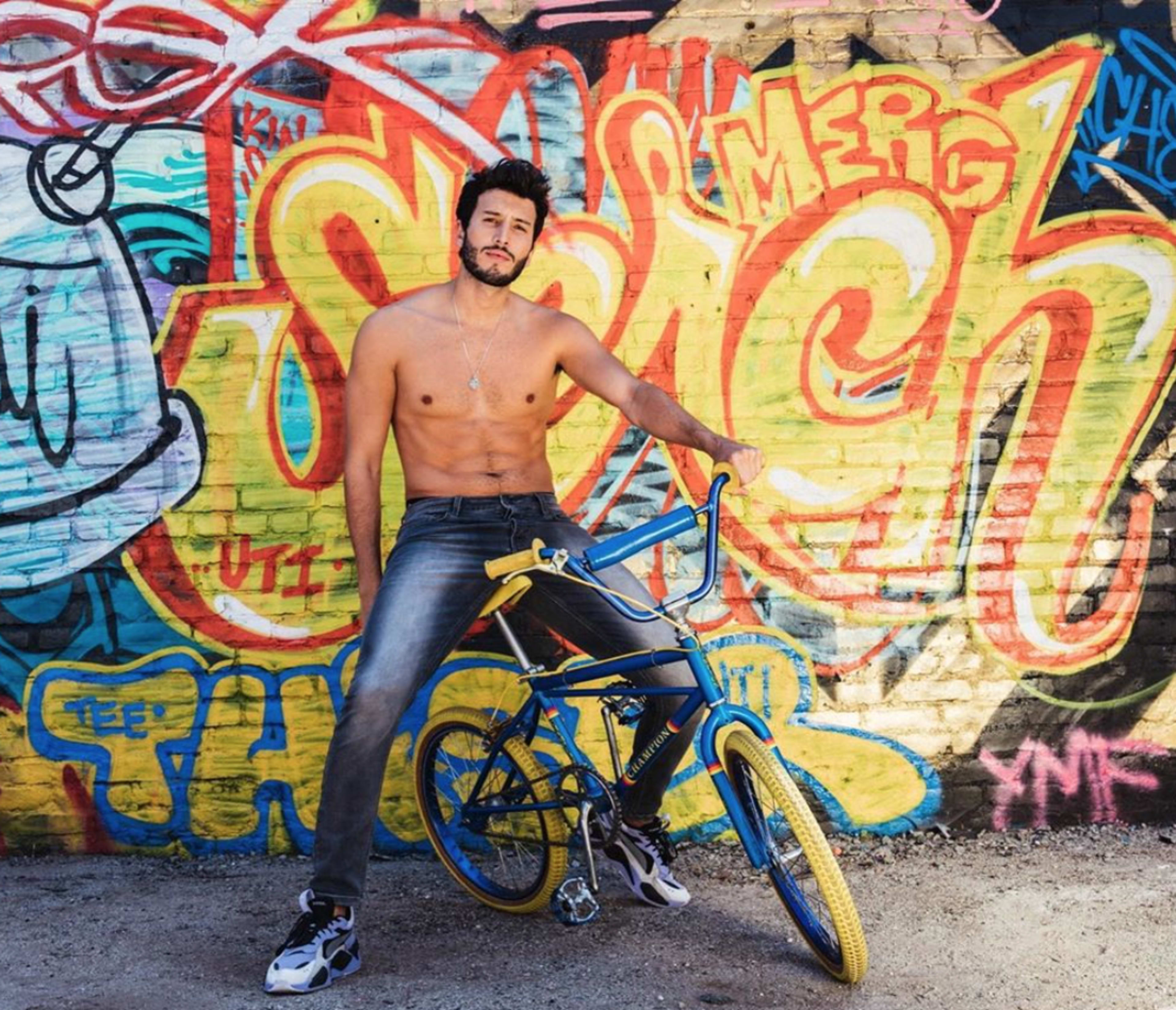 A shirtless man poses next to a blue and yellow bike in front of a vibrant graffiti mural.