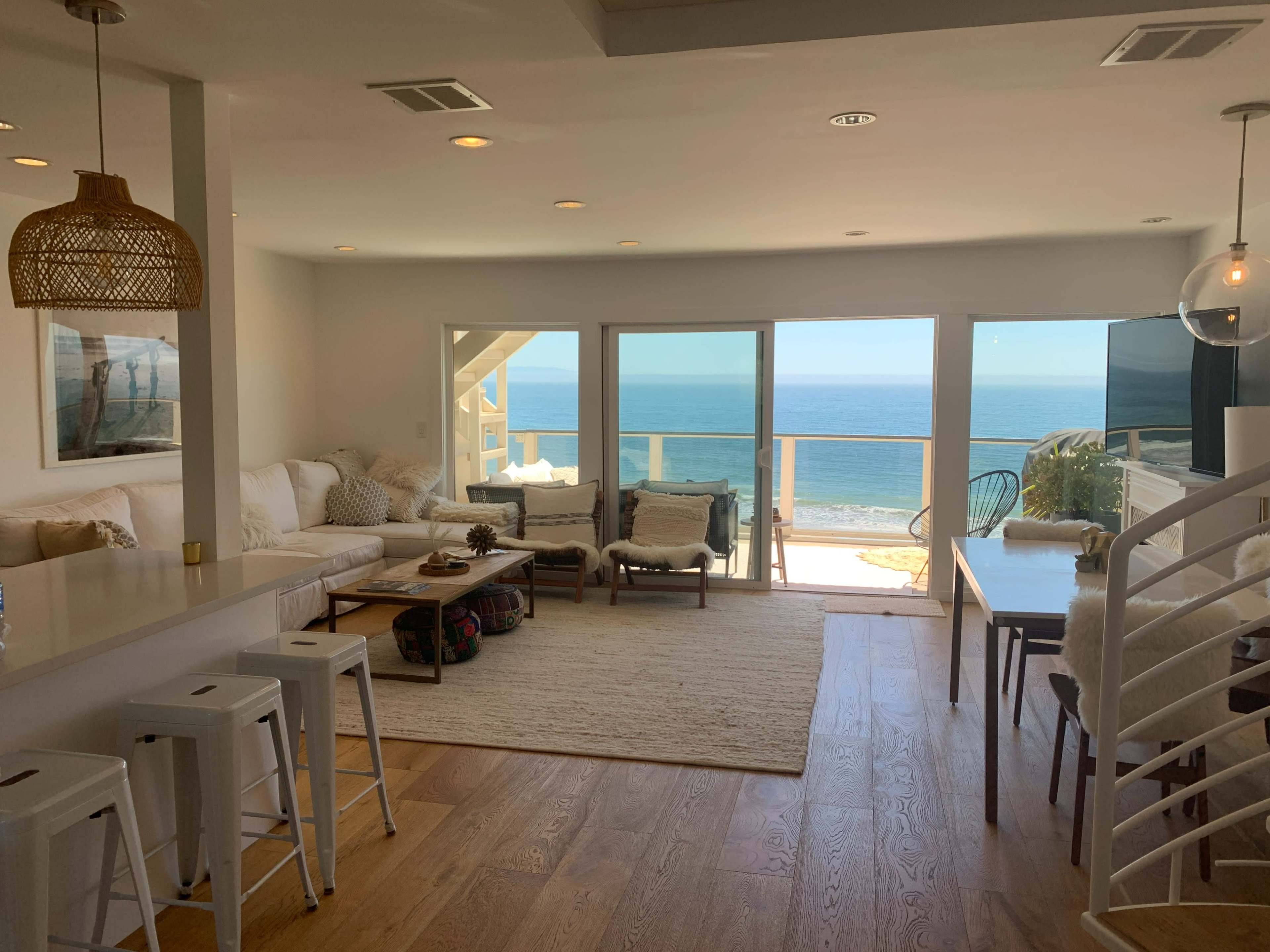 The scene depicts a coastal living space featuring a cozy seating area with a view of the ocean through large glass doors.