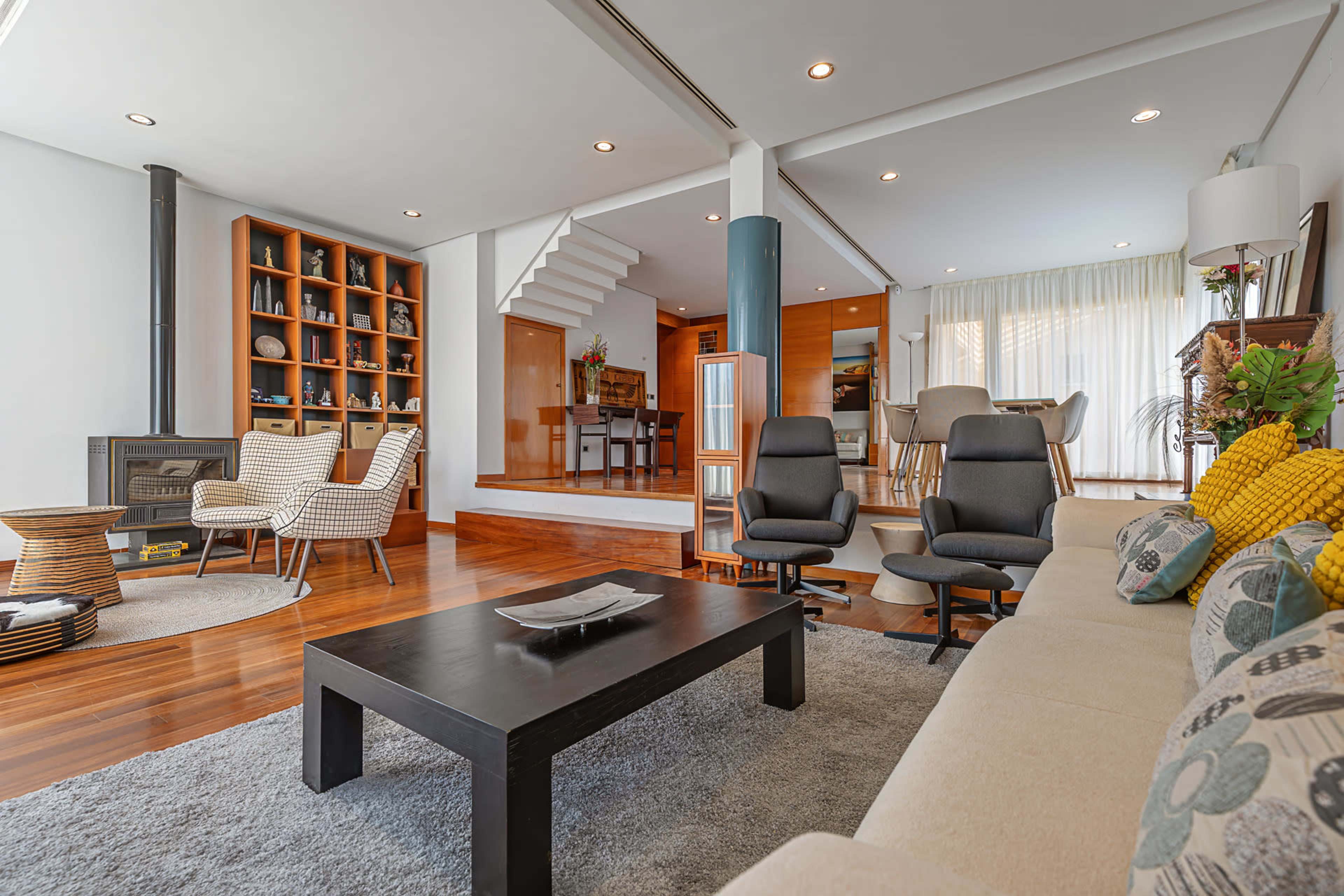 The image shows a modern living room with a sectional sofa, two armchairs, a coffee table, a bookshelf, and a staircase leading to an upper level.