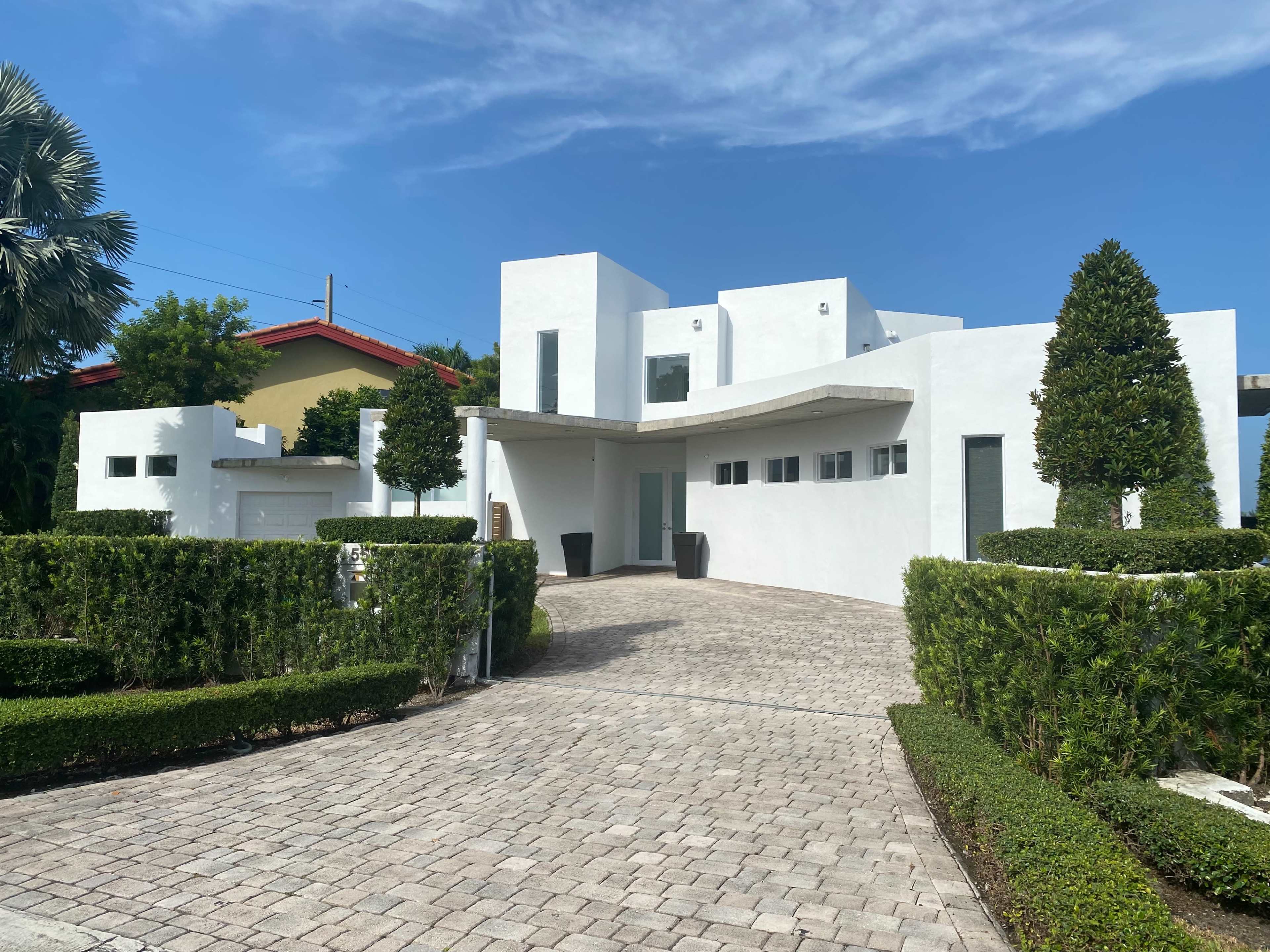 The image shows a modern white house with a unique architectural design, surrounded by neatly trimmed hedges and a paved driveway.