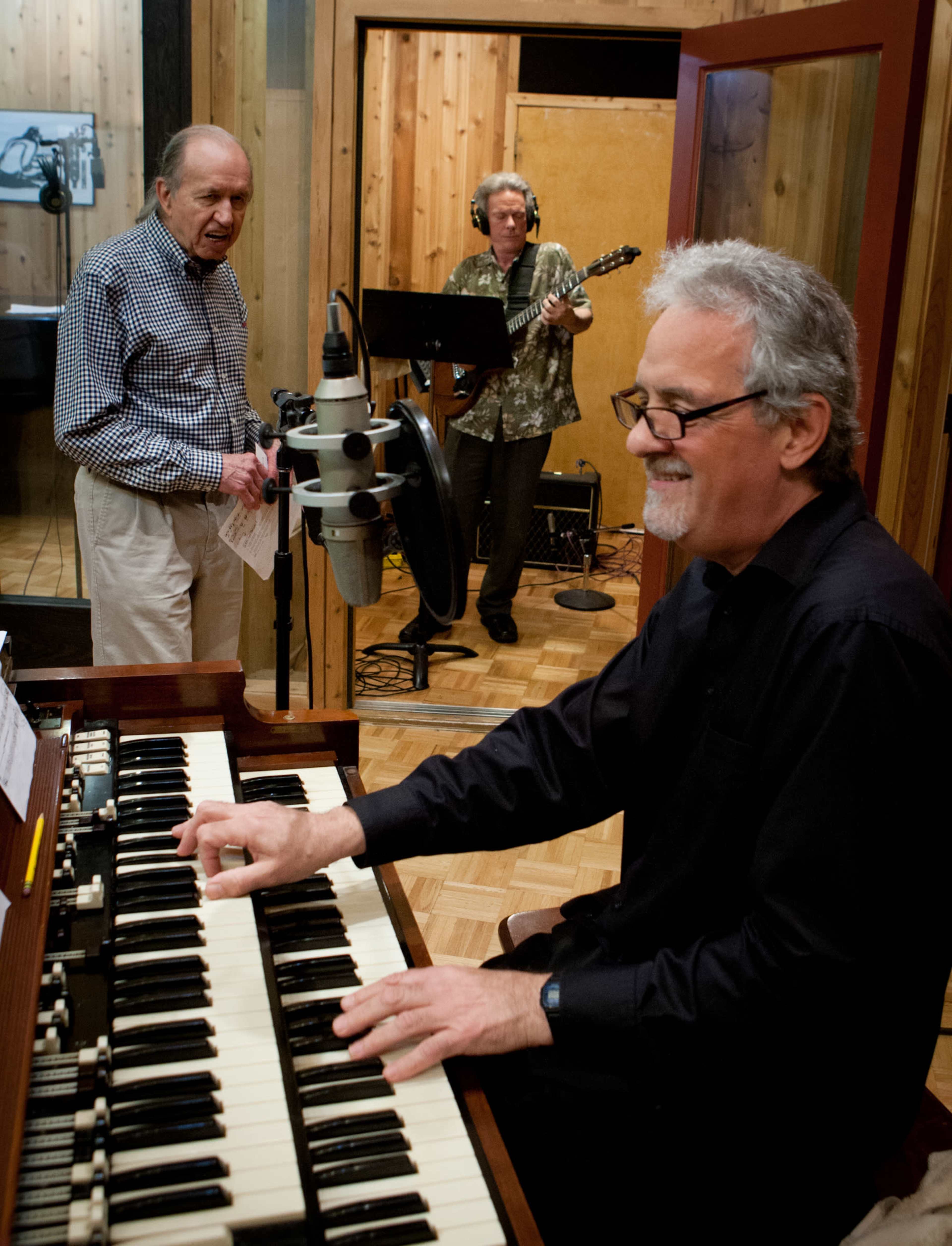 A man plays a keyboard while another man sings and a third man plays guitar in a recording studio.