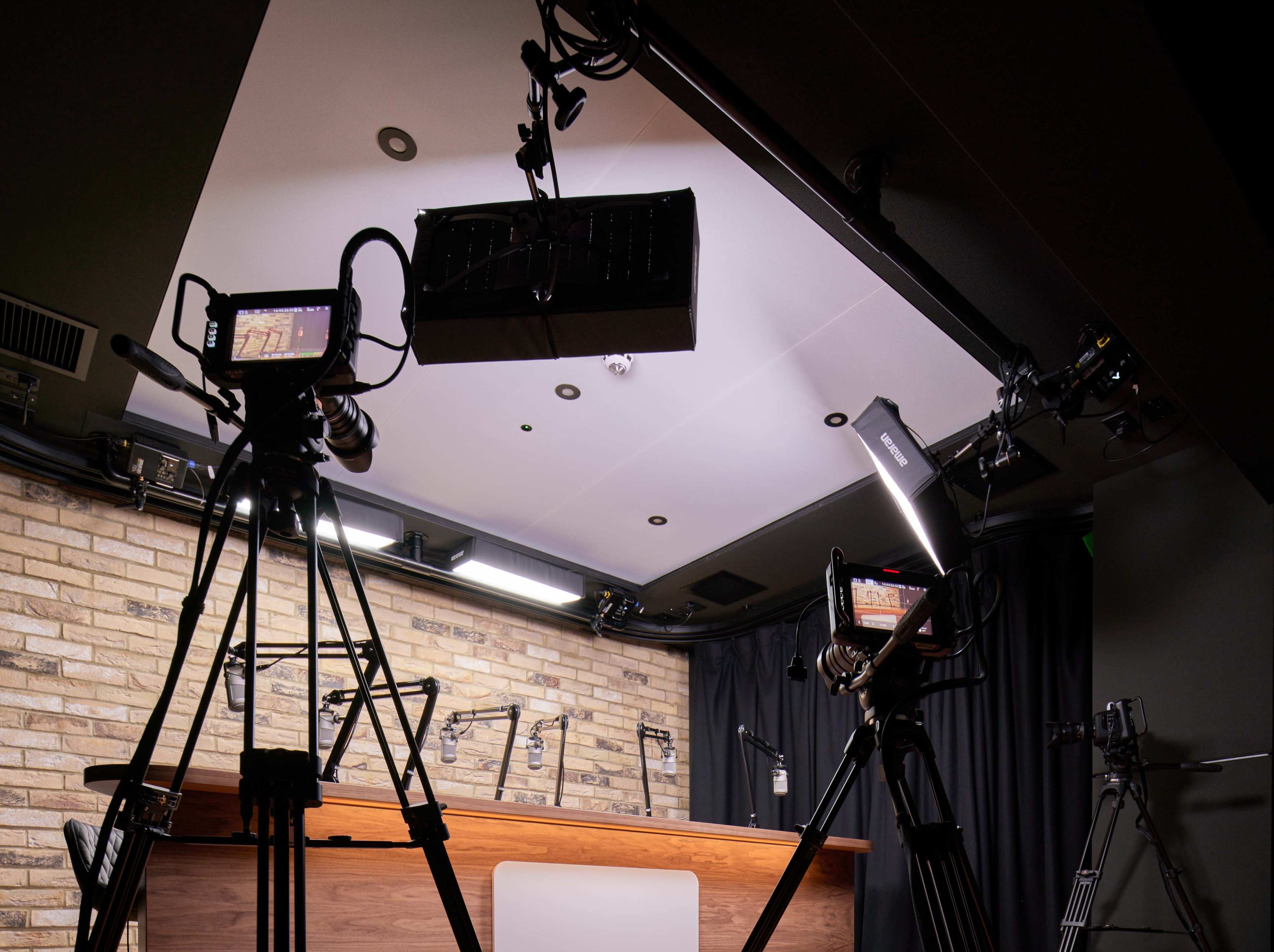 The image shows a TV studio setup with multiple cameras and lighting fixtures facing a wooden desk against a brick wall.