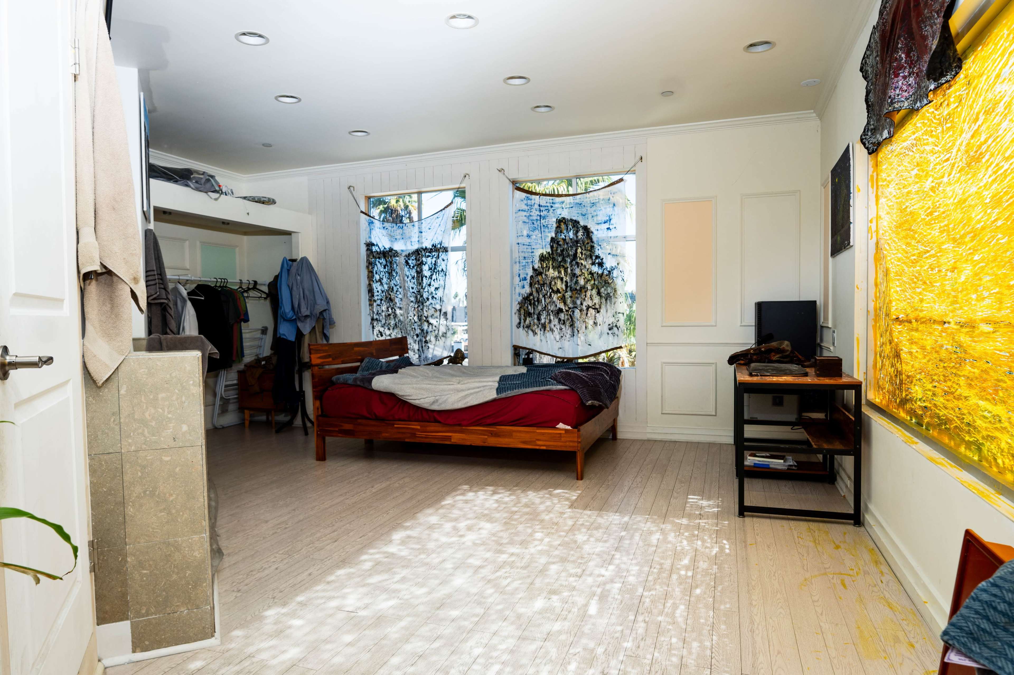 The room features a bed with a red comforter, two windows with sheer curtains, a small television, and a wardrobe area.
