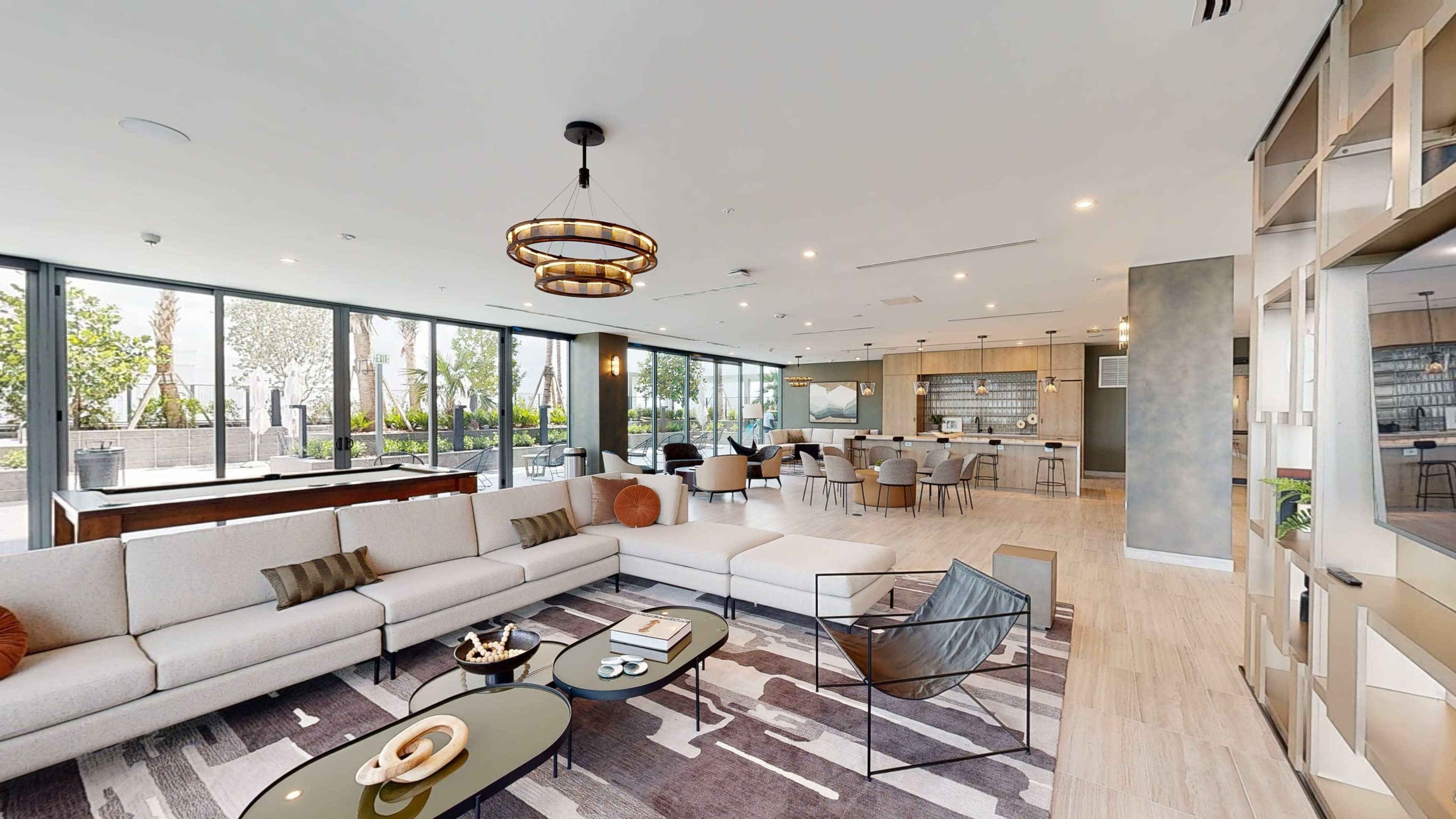 A modern common area features a spacious lounge with a sectional sofa, a coffee table, and a dining area in the background, all surrounded by large windows.