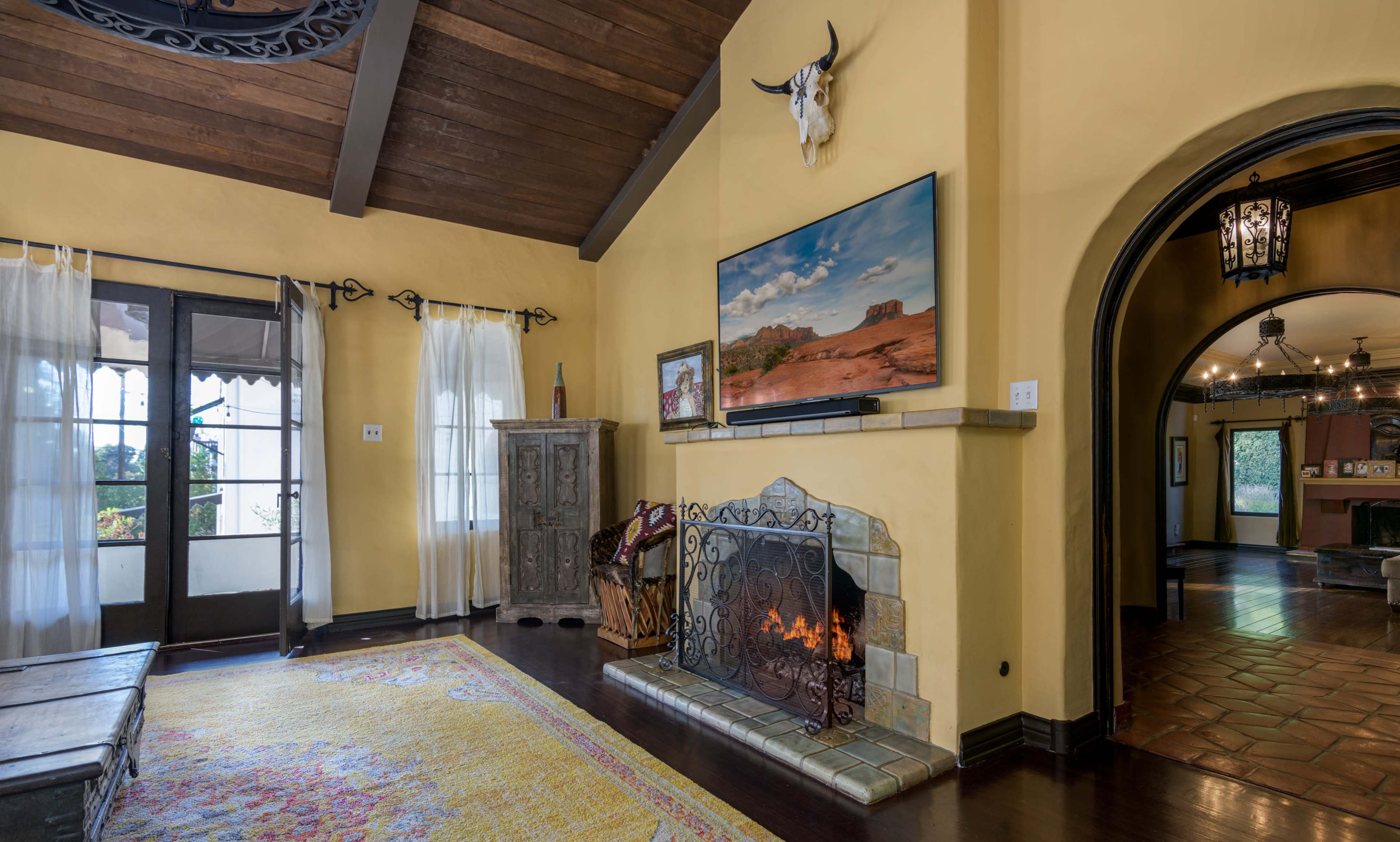 The image shows a living room featuring a fireplace with a decorative iron screen, a large television mounted above it, and arched doorways leading to other spaces.