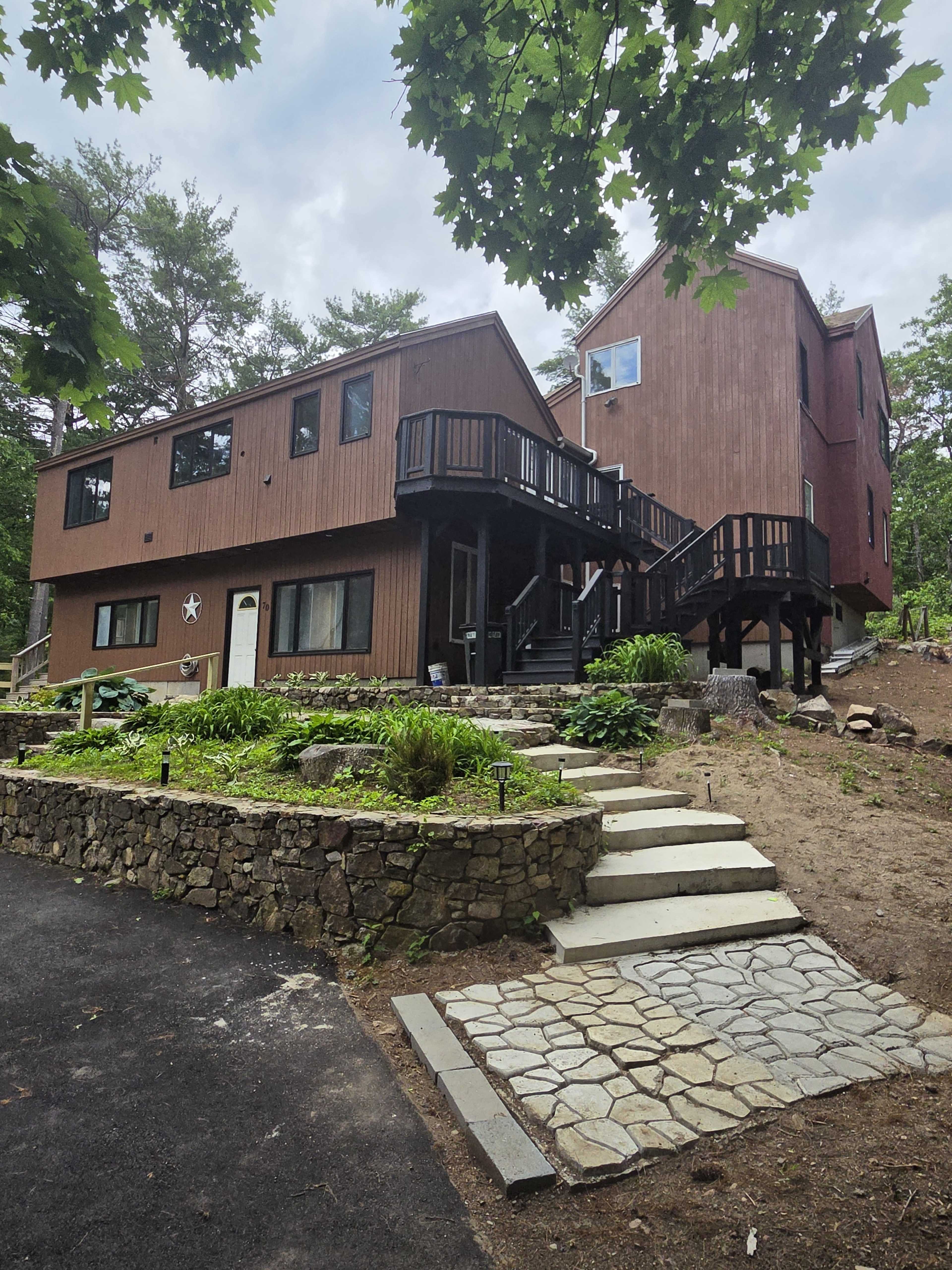 The image shows a two-story wooden house with a wrap-around deck, surrounded by trees and a stone pathway leading up to the entrance.