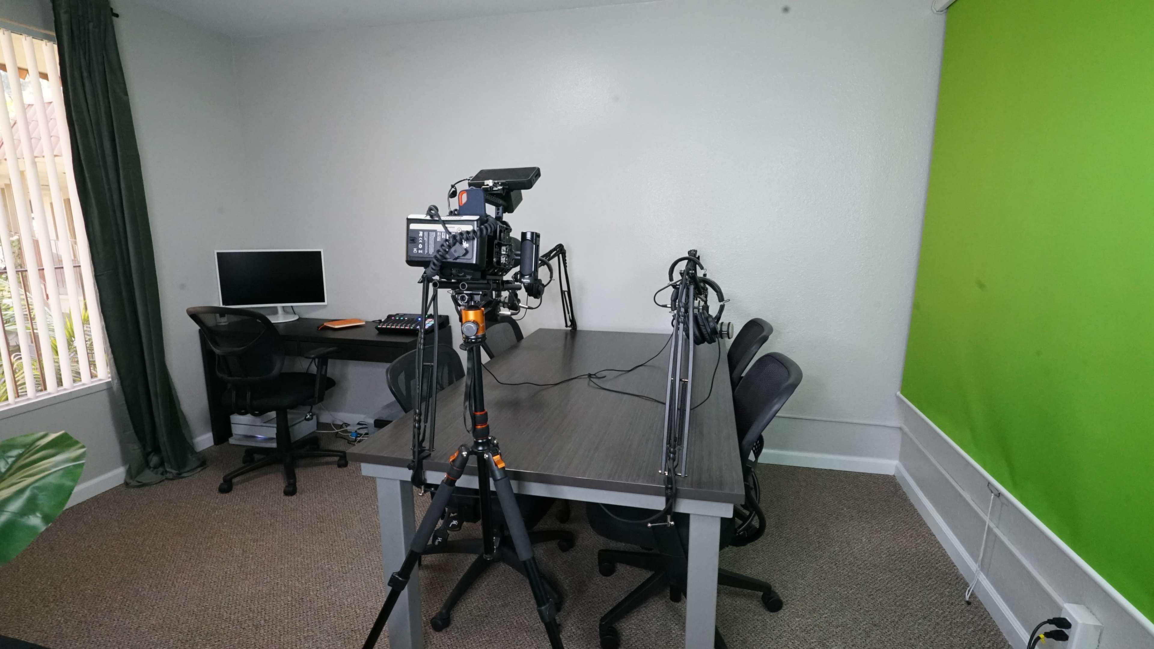 A camera is set up on a tripod facing a table with three chairs in a room featuring a green wall, a computer, and a window with blinds.
