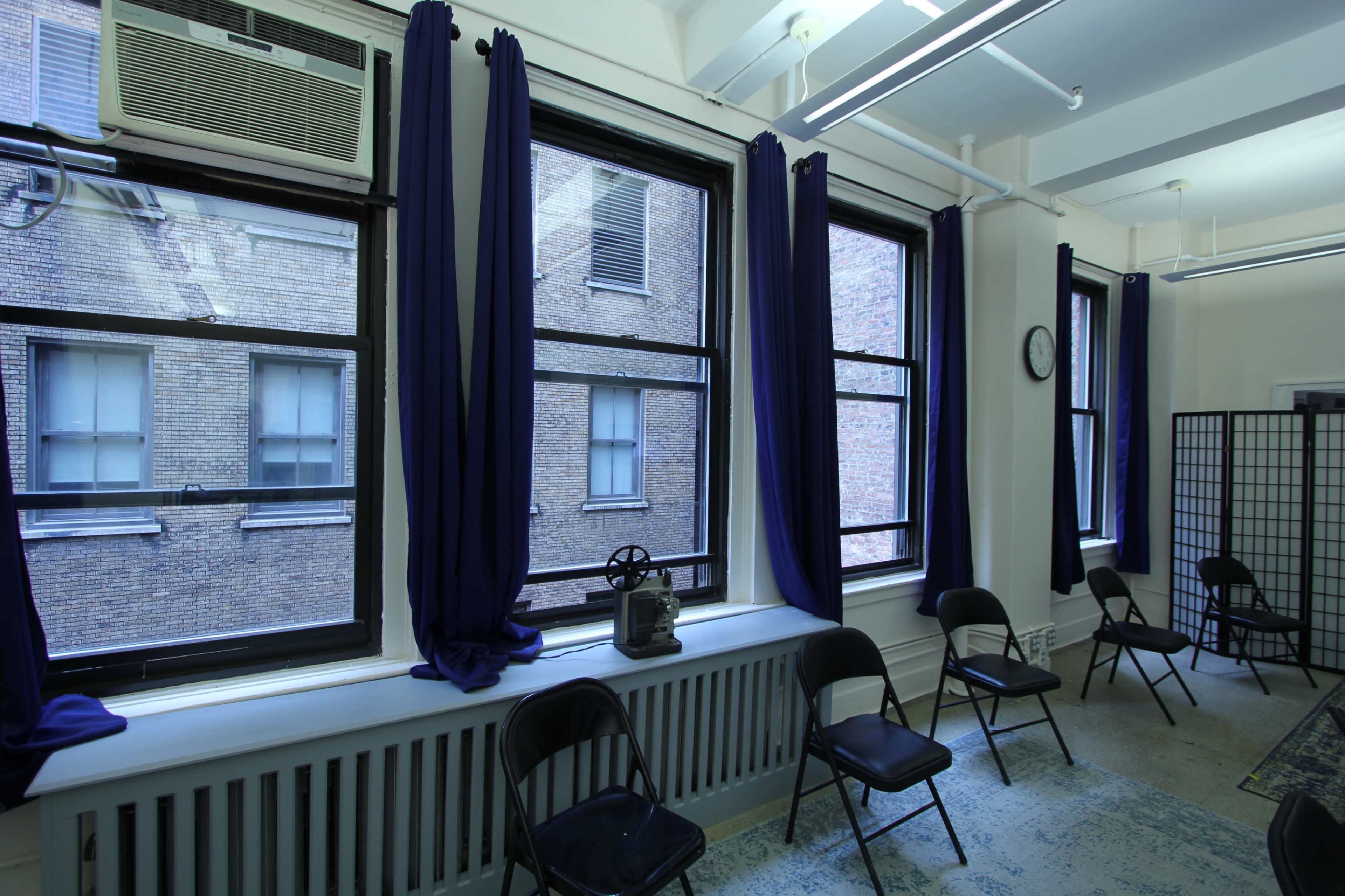 The image shows an empty room with several black chairs arranged in front of windows covered with dark curtains, revealing a view of a brick wall outside.