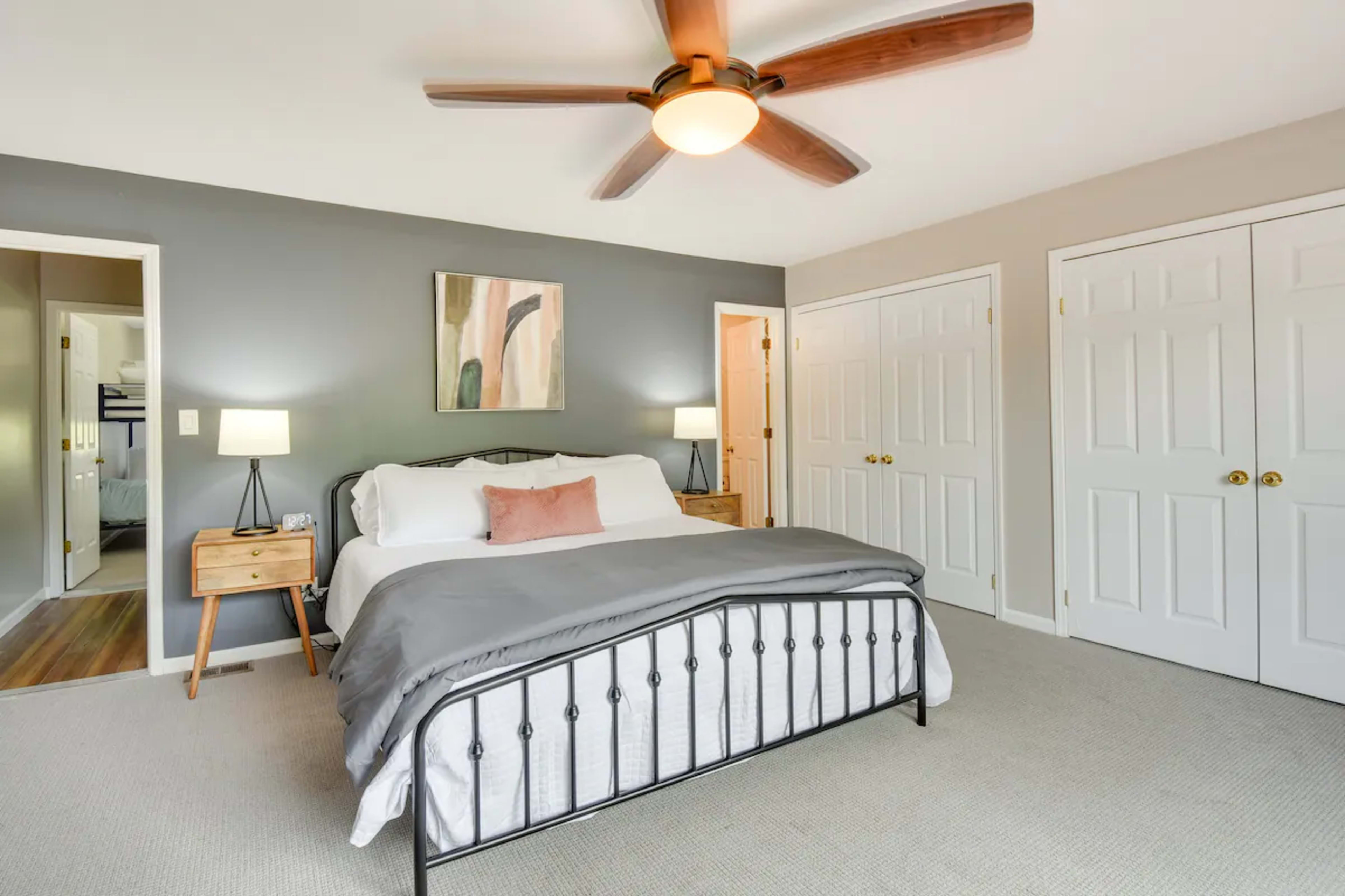 A spacious bedroom features a bed with a gray blanket and a ceiling fan, with two doors leading to other areas and a painting on the wall.