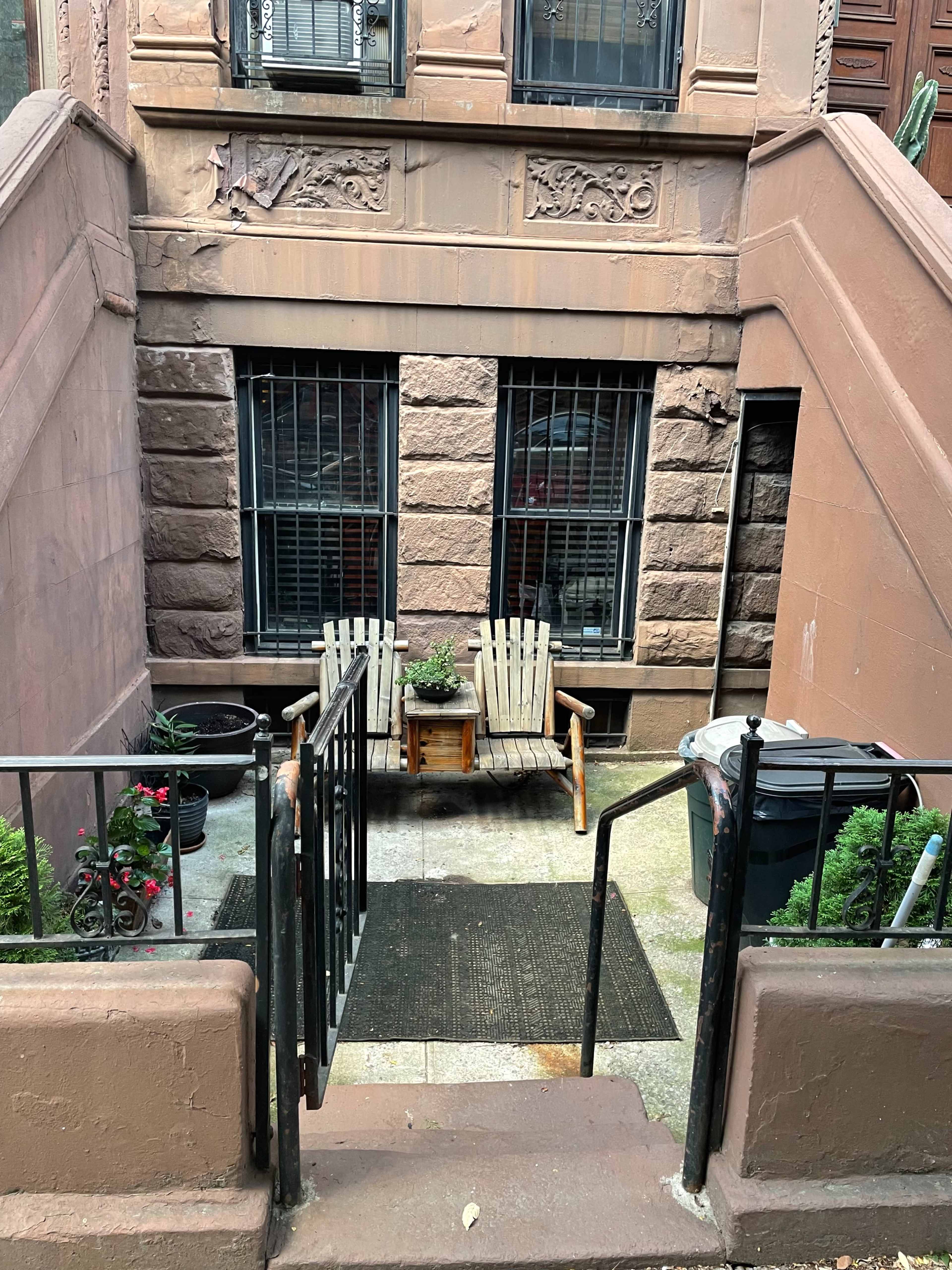 The image shows a small outdoor space between two brownstone buildings with two wooden chairs, a table with a plant, and a trash bin.