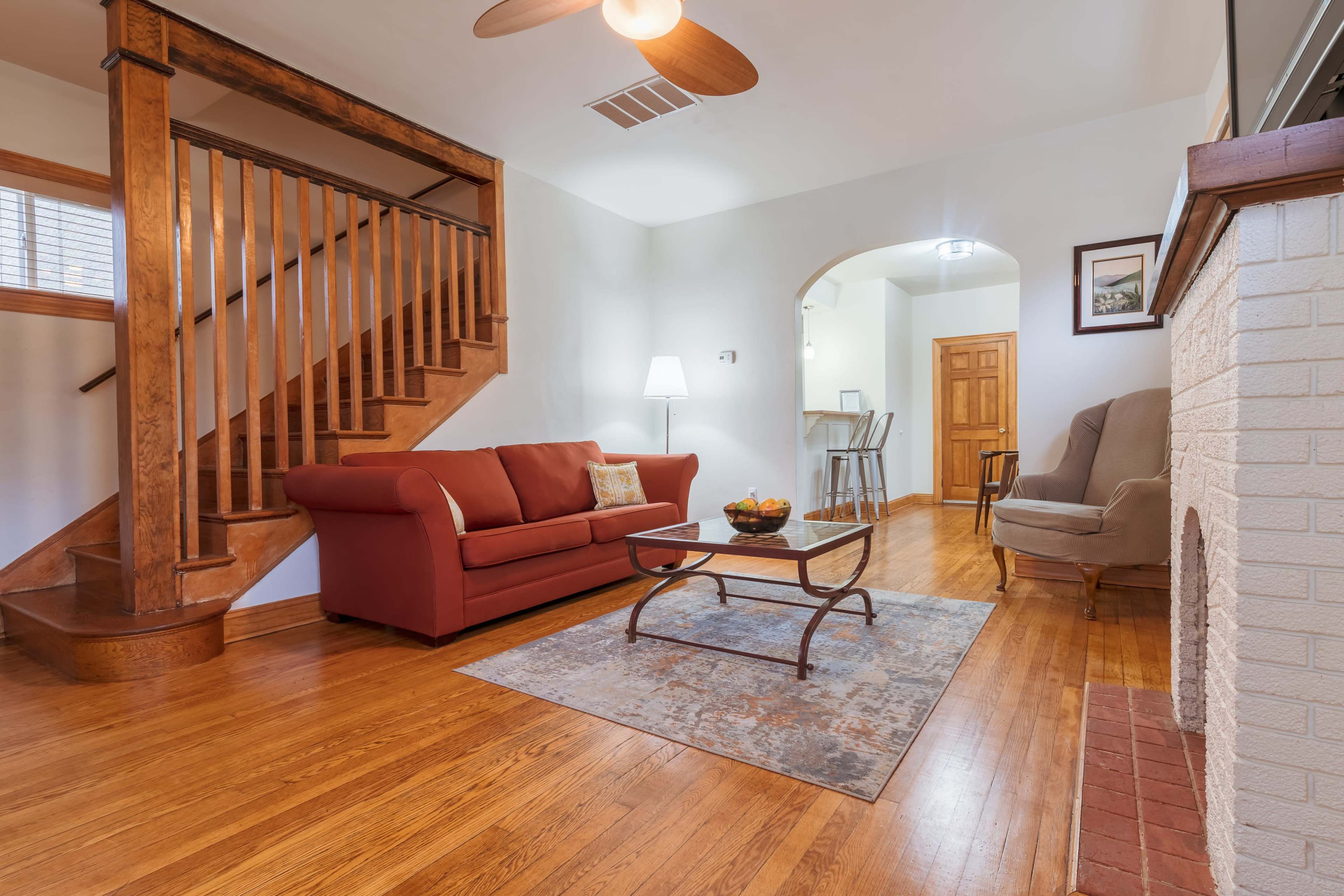 A cozy living room features a red sofa, a coffee table with a bowl of fruit, and a staircase leading to the upper level.