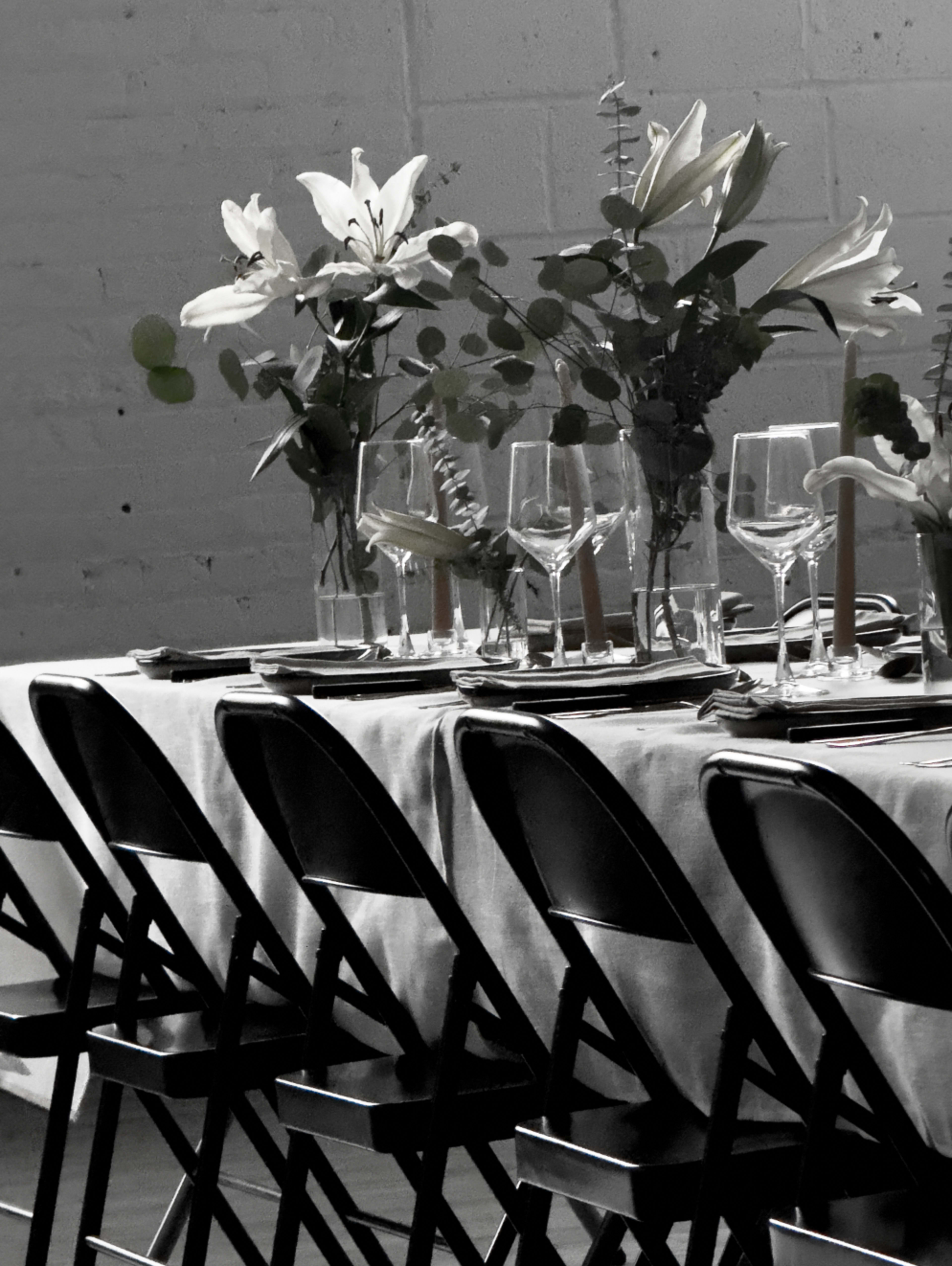 A long table set for a gathering is adorned with white flowers and glassware, lined with black folding chairs.