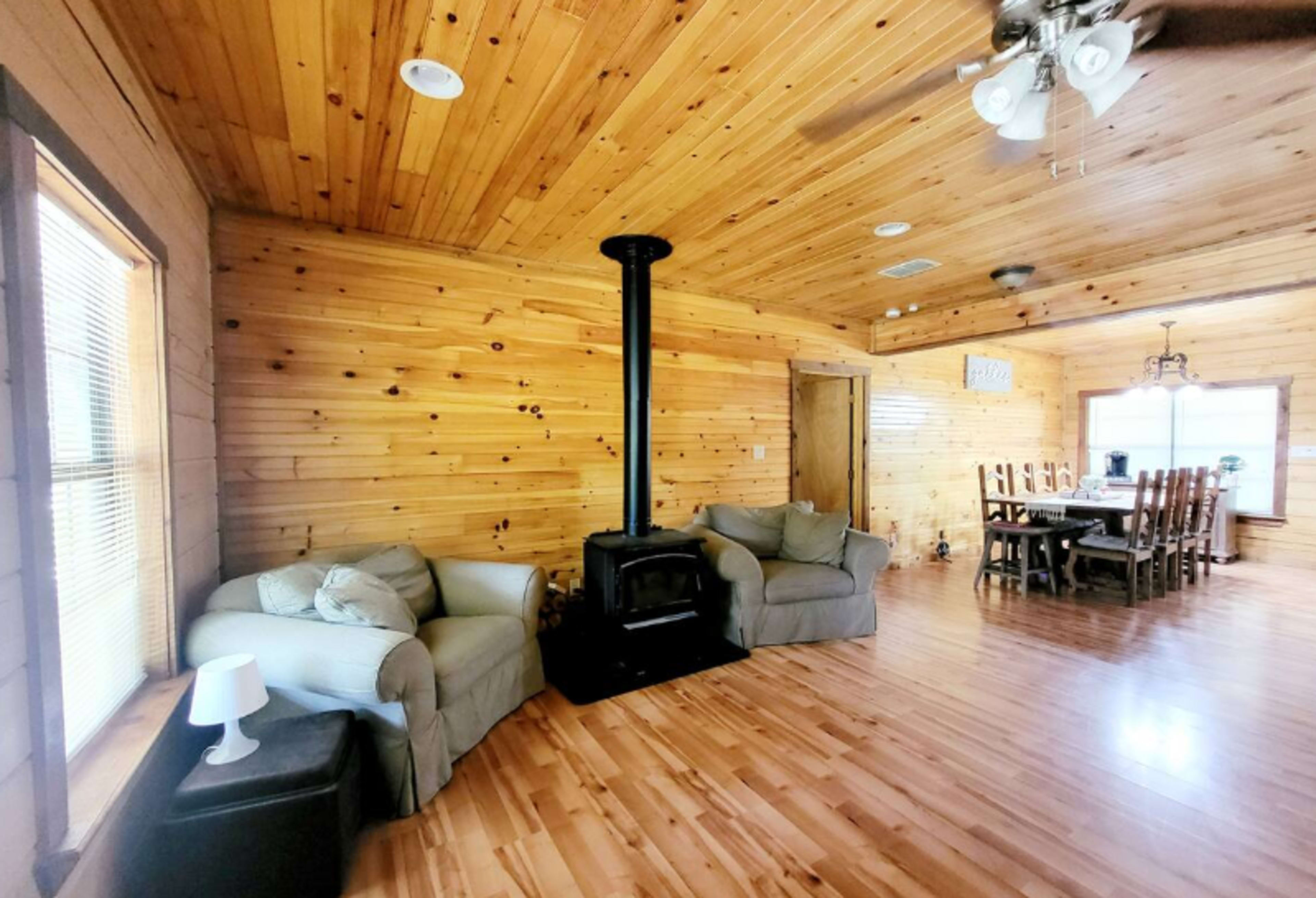 The image shows a warm wooden living space with a wood stove, two armchairs in a corner, and a dining table with chairs in the background.