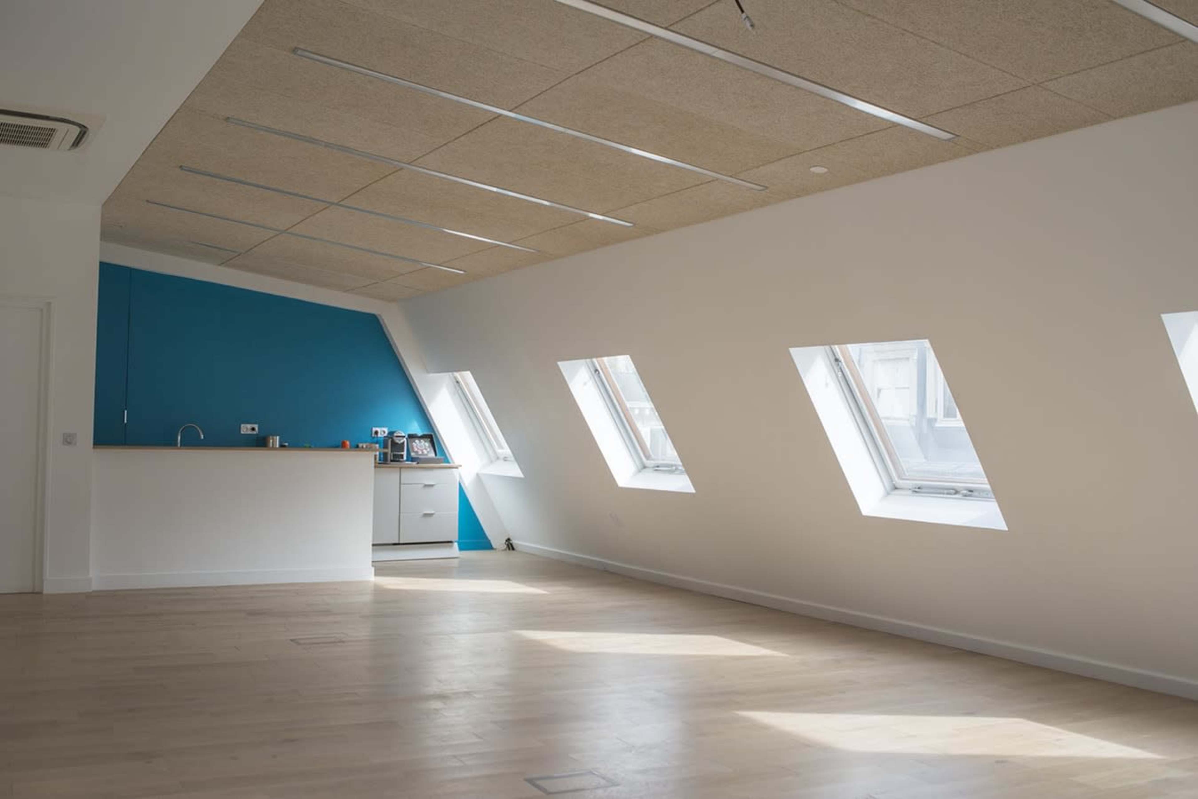A spacious, well-lit room with angled windows and a blue accent wall, featuring a small kitchenette along one side.