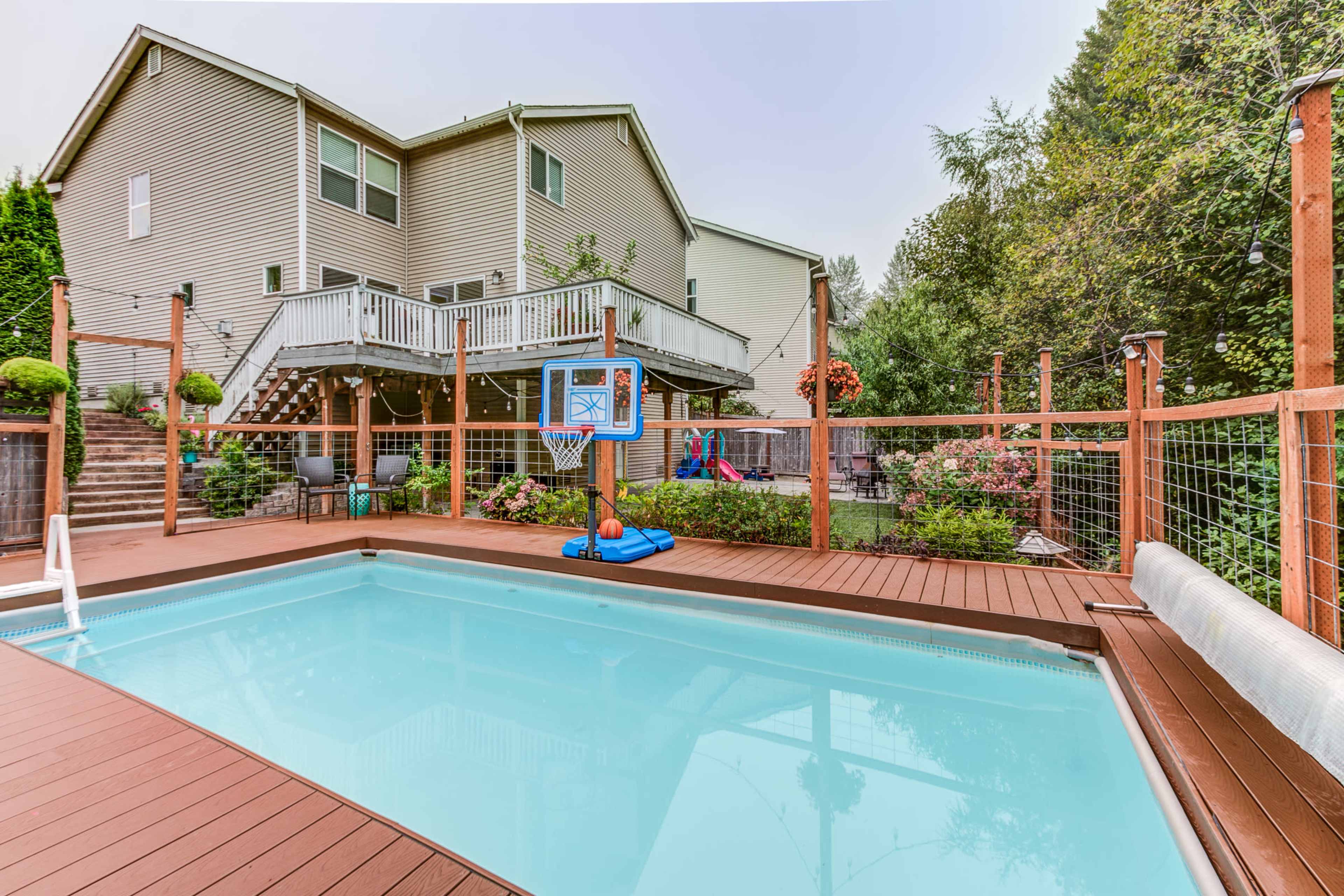 A backyard features a swimming pool surrounded by a wooden deck and landscaped area, with a house and children's play equipment visible in the background.