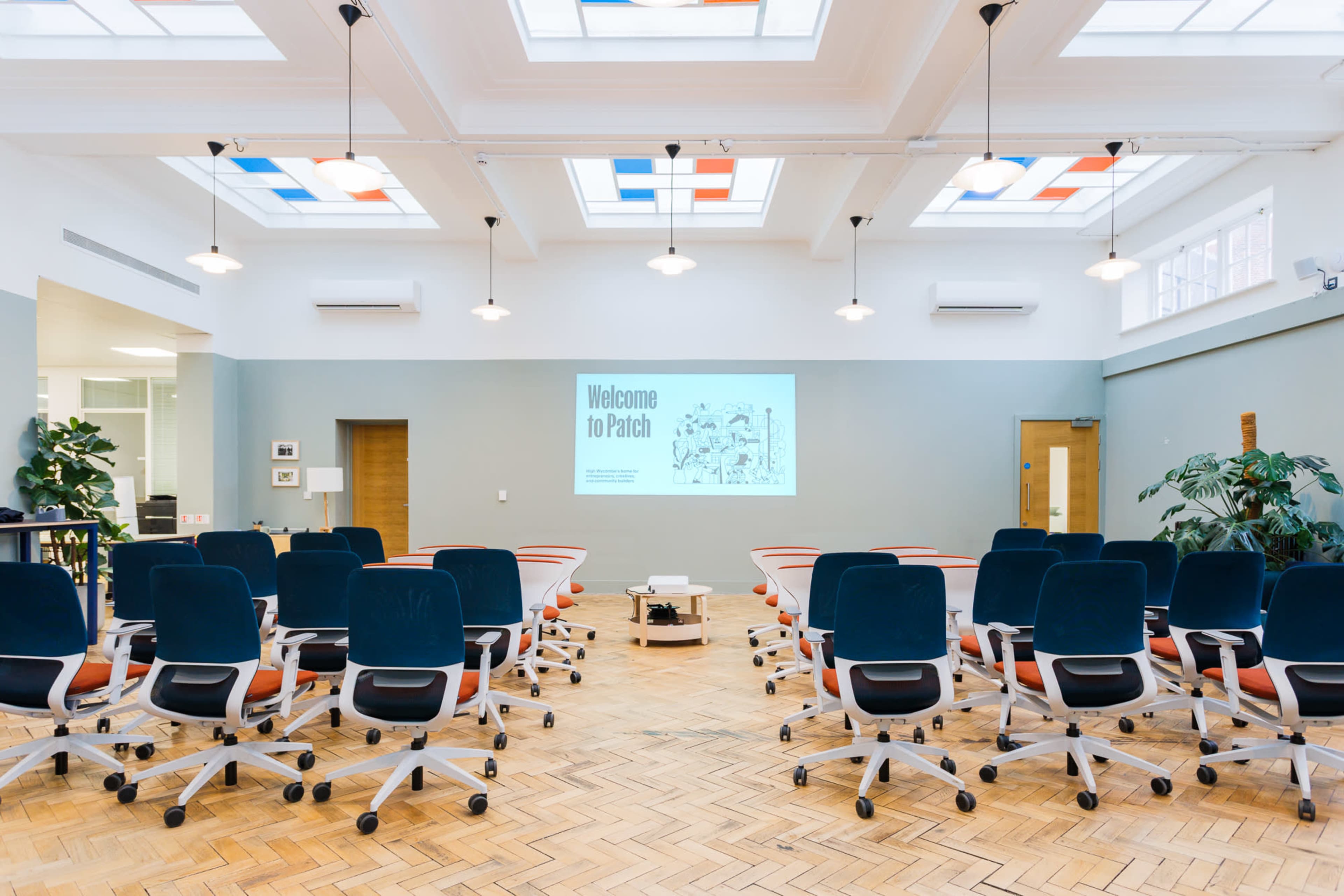 A spacious conference room features rows of blue and orange office chairs facing a large projection screen on a pastel wall, with skylights above providing natural light.