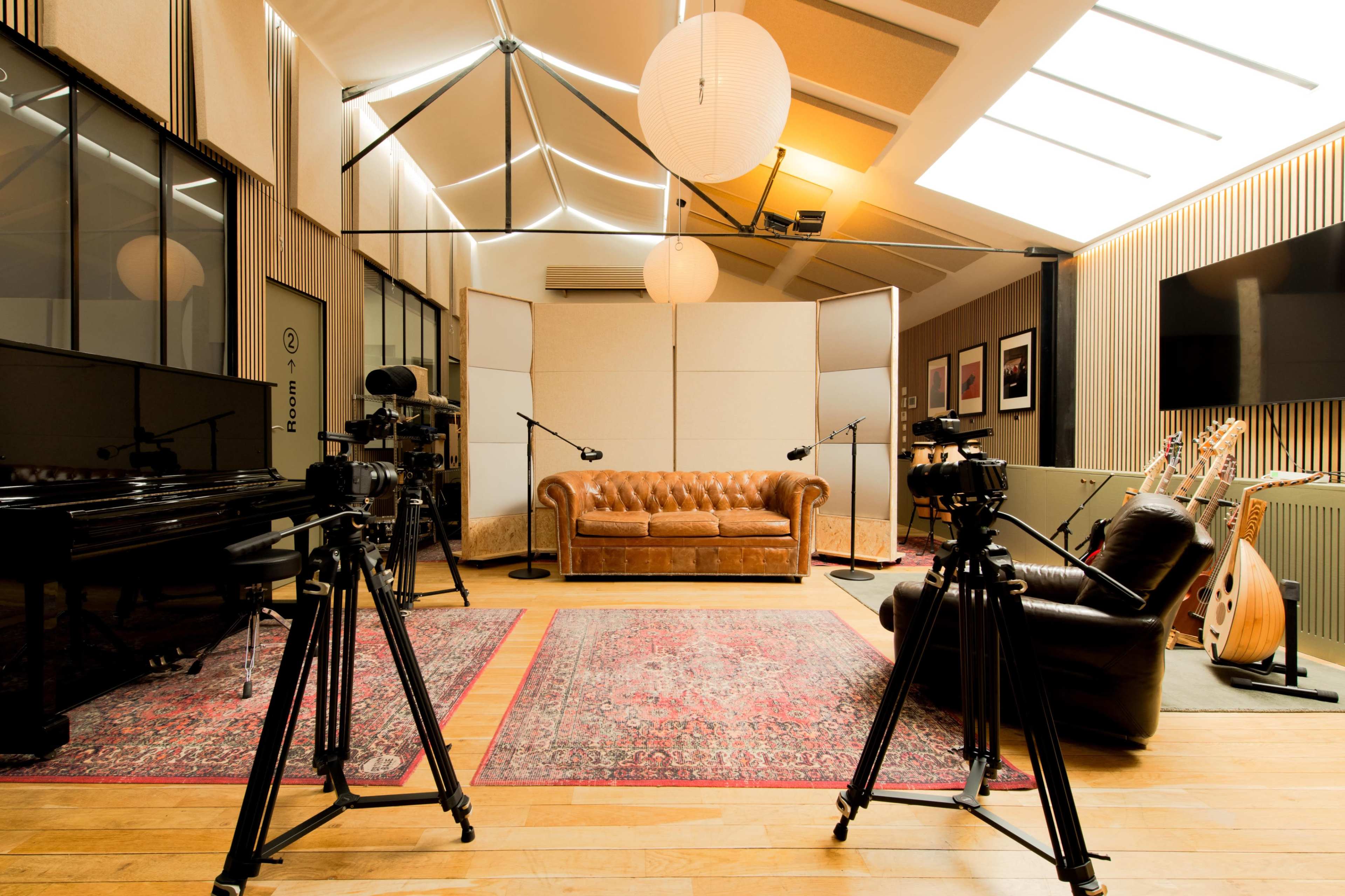 The scene features a studio with a plush brown leather couch, two cameras on tripods facing the seating area, a grand piano, and various musical instruments displayed against a textured wall.
