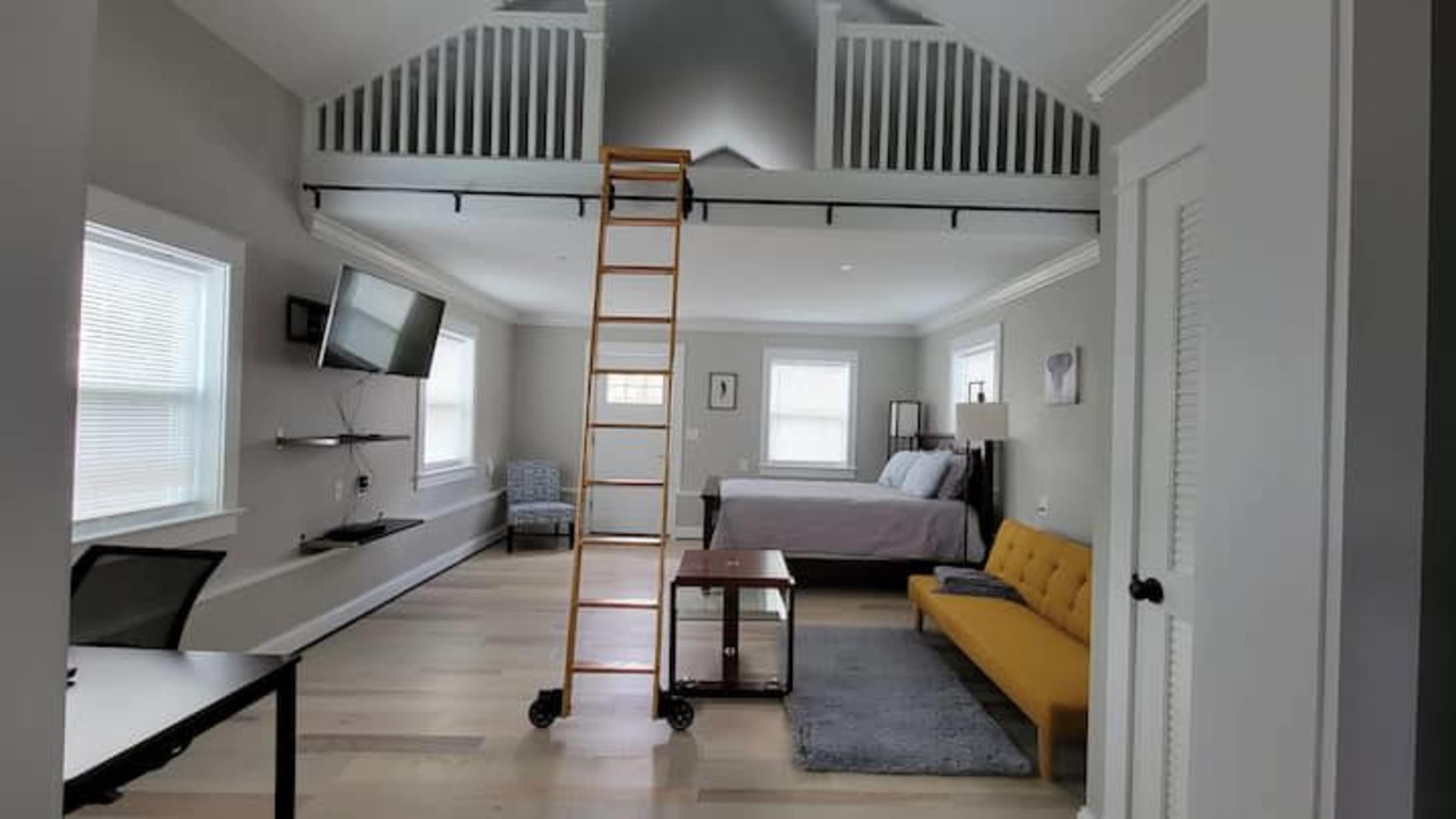 The image shows a modern room with a lofted area, featuring a bed, a yellow sofa, a desk, and a ladder leading to the upper level.
