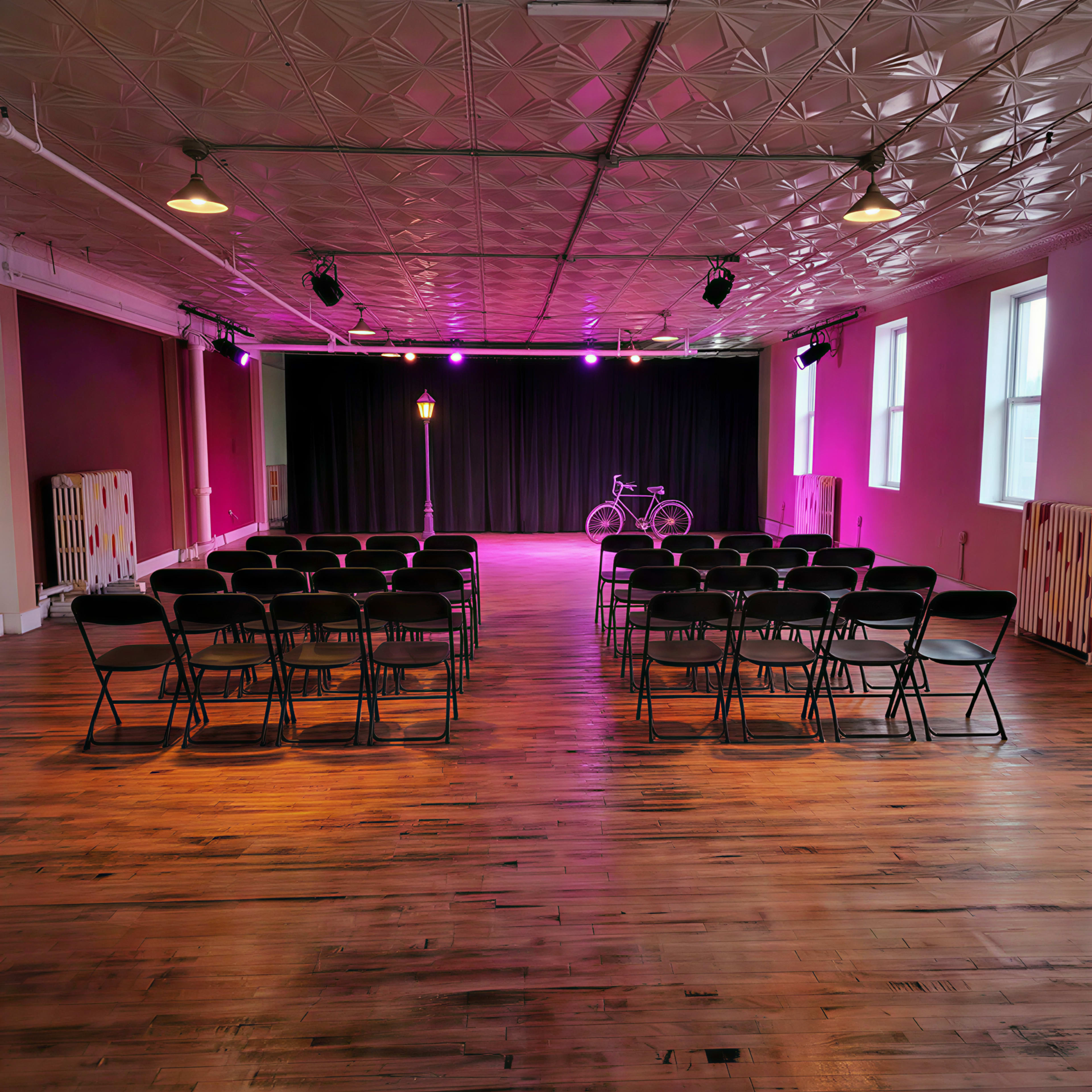 The image shows a spacious performance venue with rows of black chairs facing a stage illuminated by purple lighting, featuring a bike and a lamppost on one side.