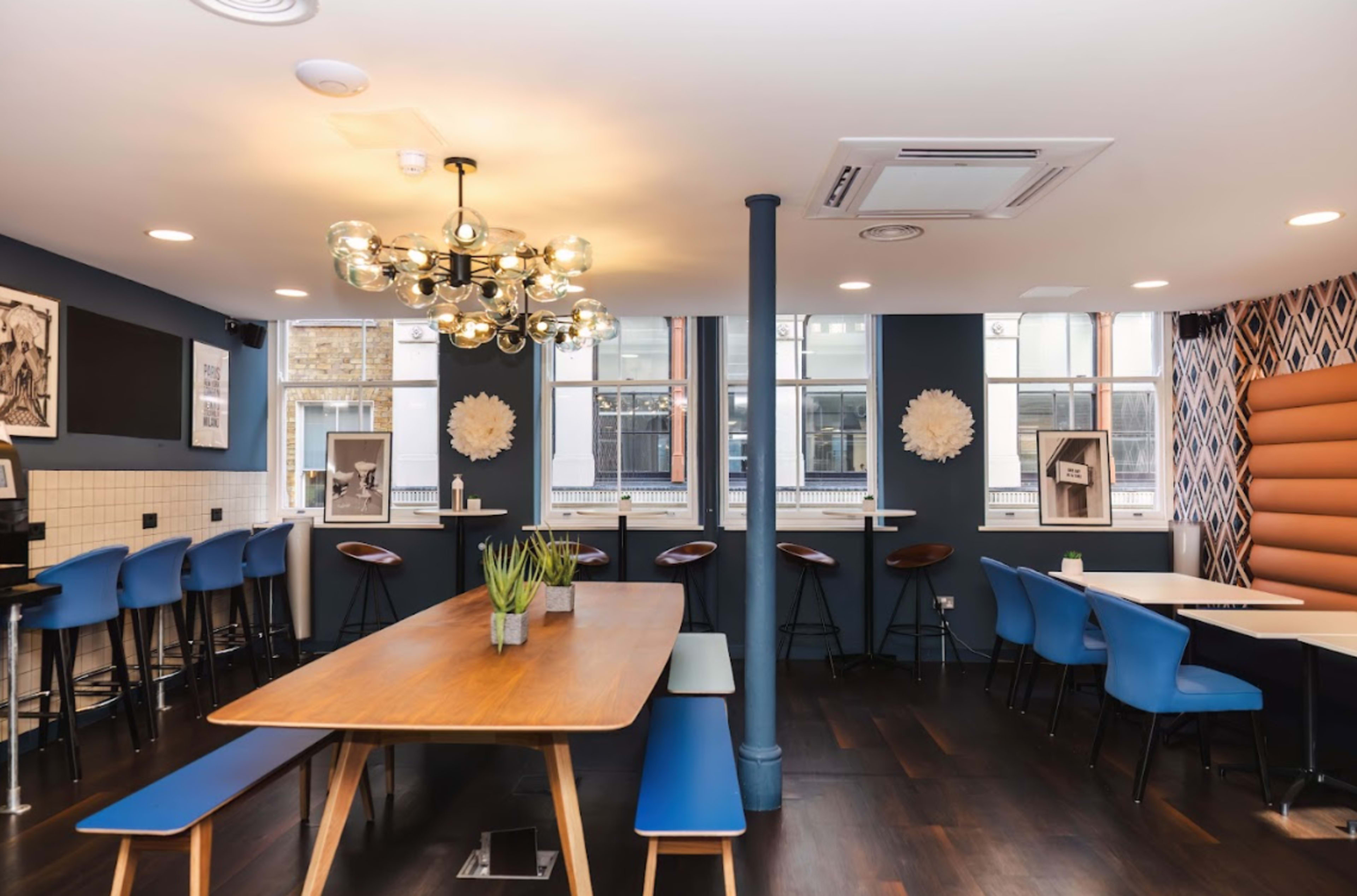 A modern café interior features a long wooden table surrounded by blue chairs and a hanging light fixture, with large windows providing natural light.