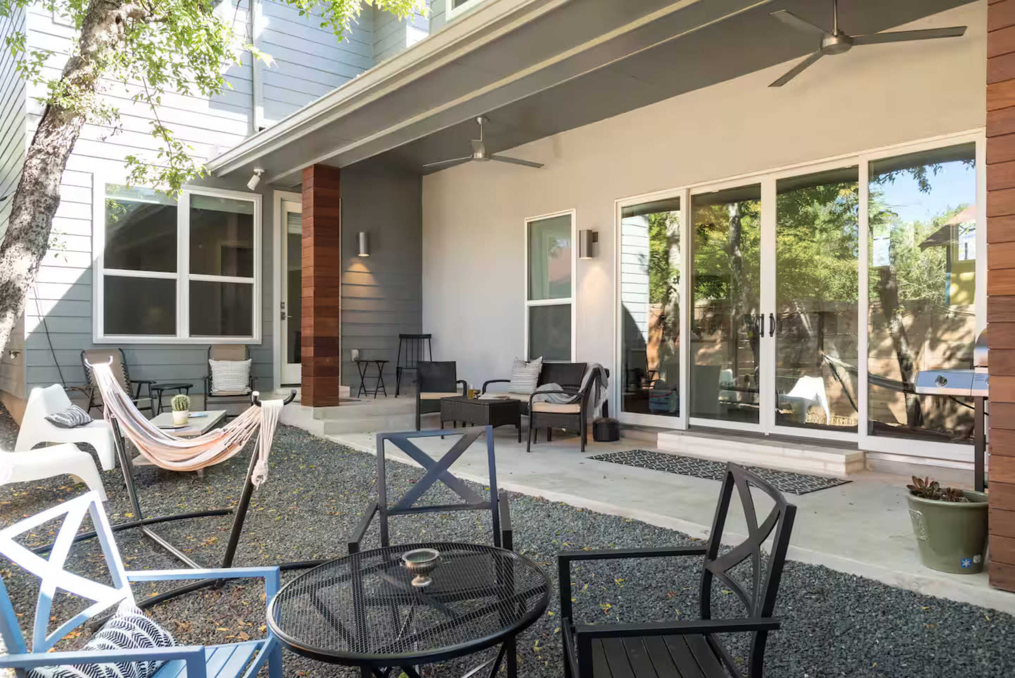 The image shows a backyard patio with seating areas comprised of chairs, a hammock, and a coffee table, surrounded by gravel and trees.
