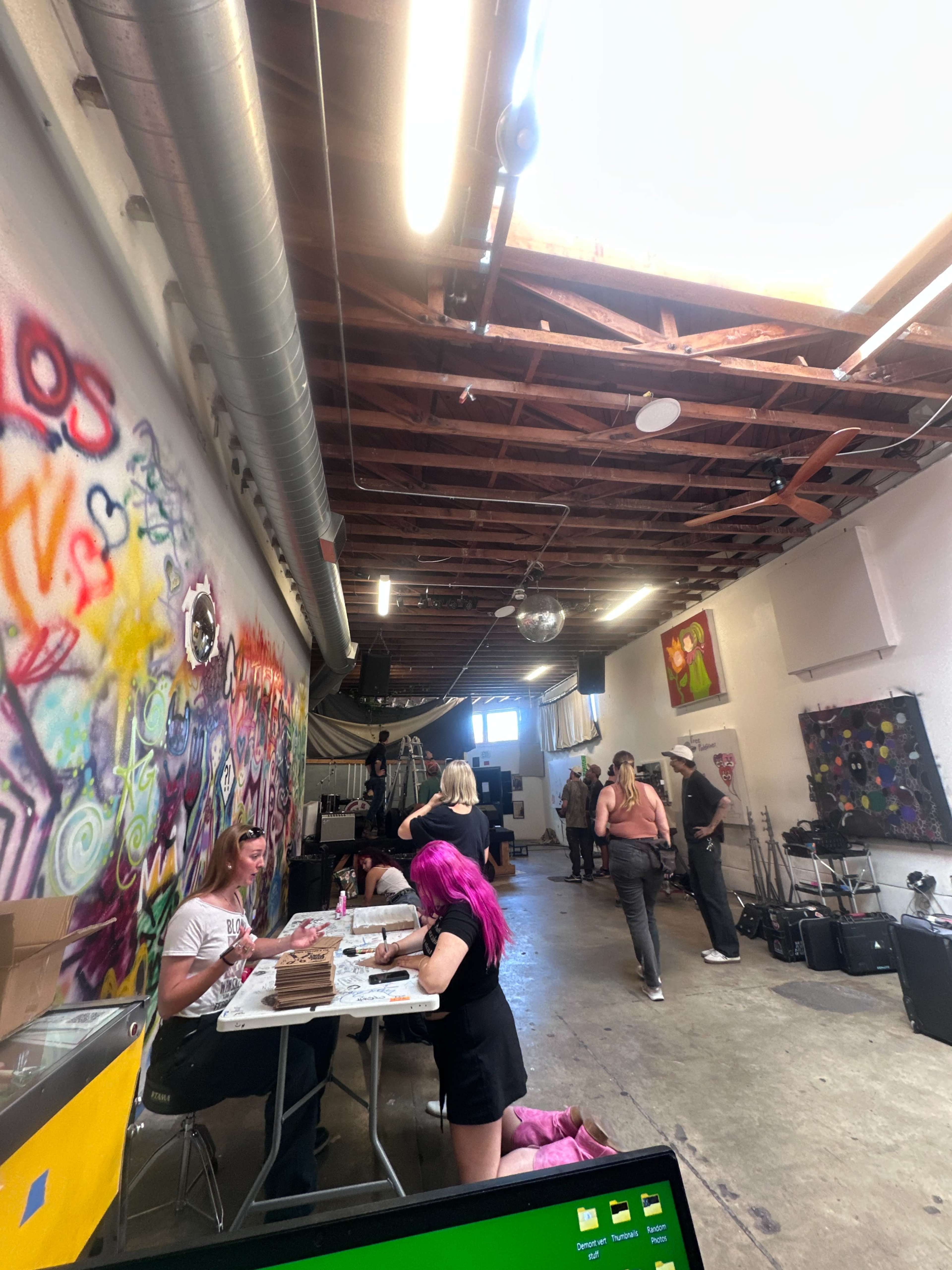The image shows a lively, graffiti-decorated studio space with various people engaged in activities, including crafting and socializing, under bright overhead lighting.