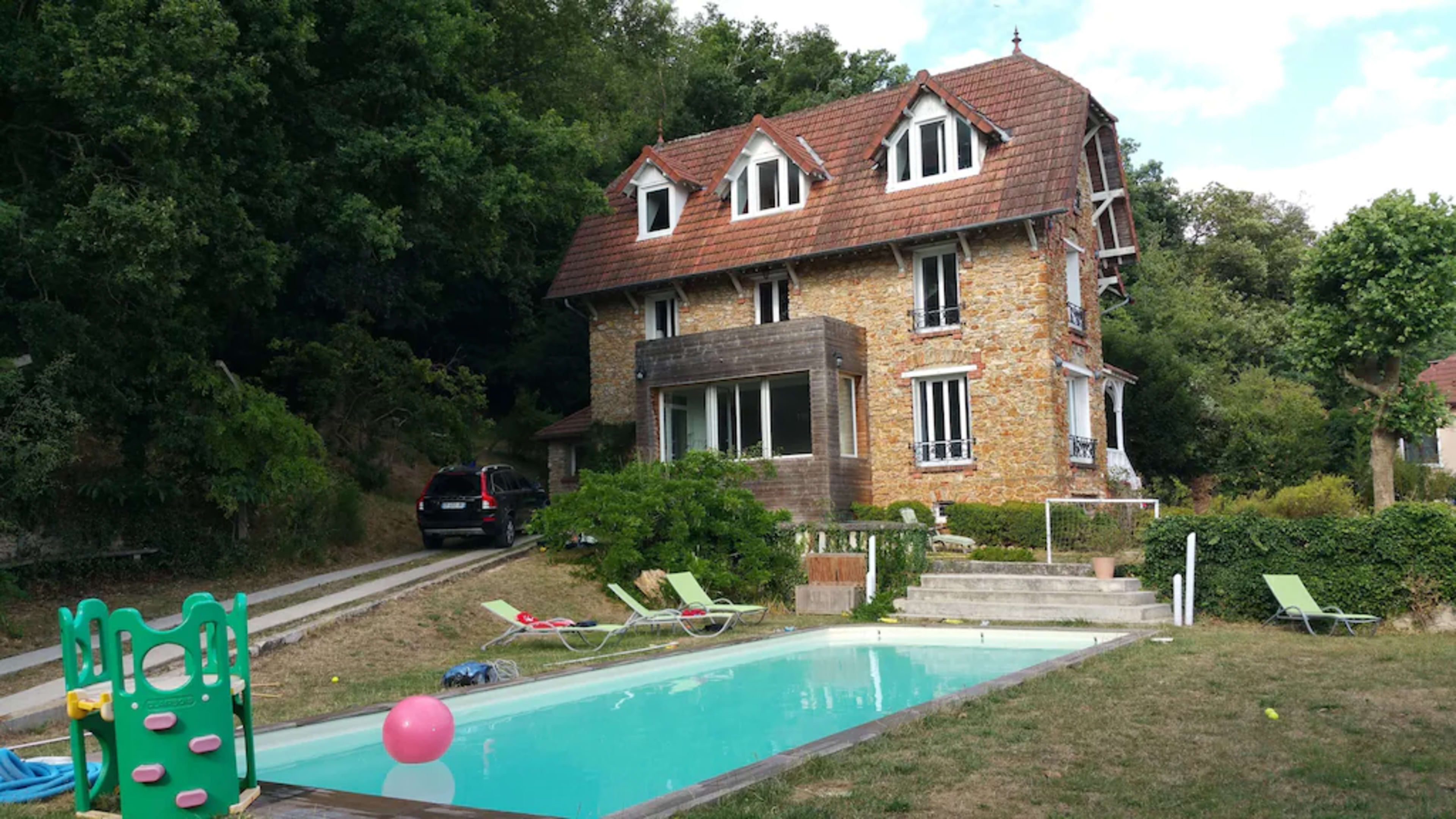 A three-story stone house with a sloped roof is situated near a swimming pool and surrounded by greenery.