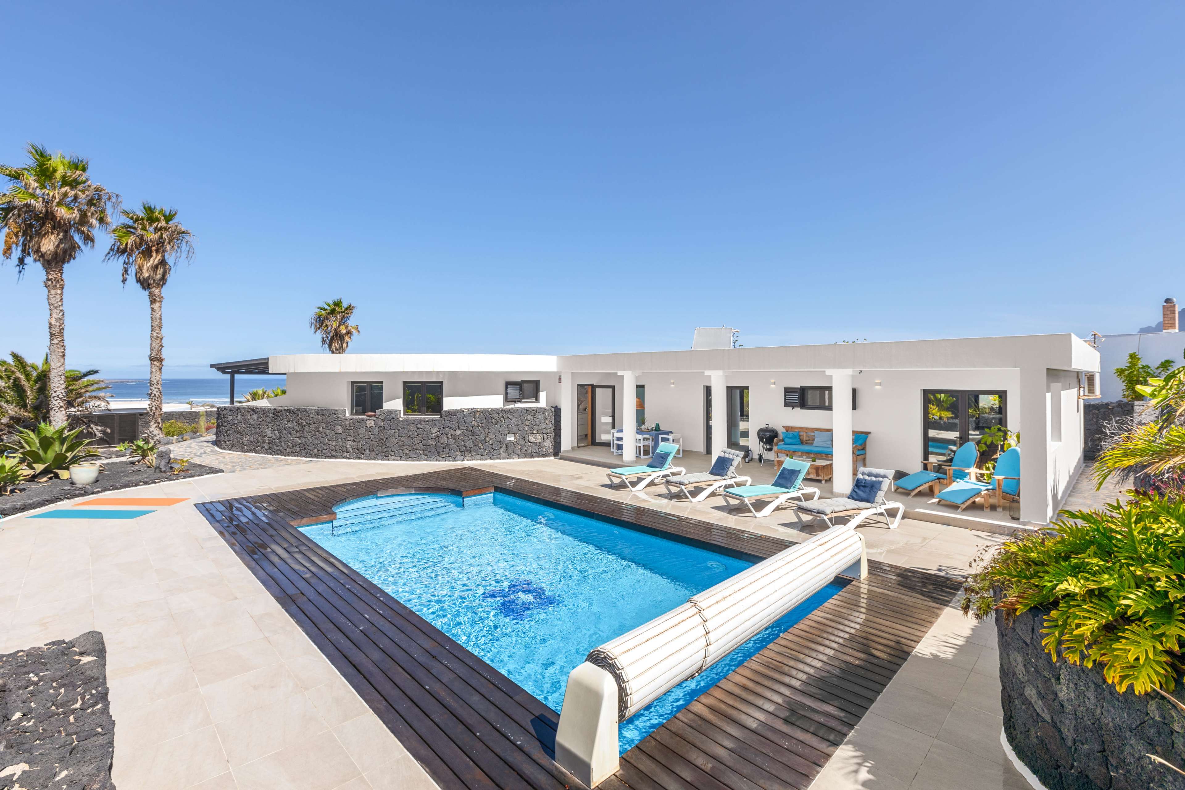The image shows a modern white villa with a swimming pool, surrounded by palm trees and outdoor seating.