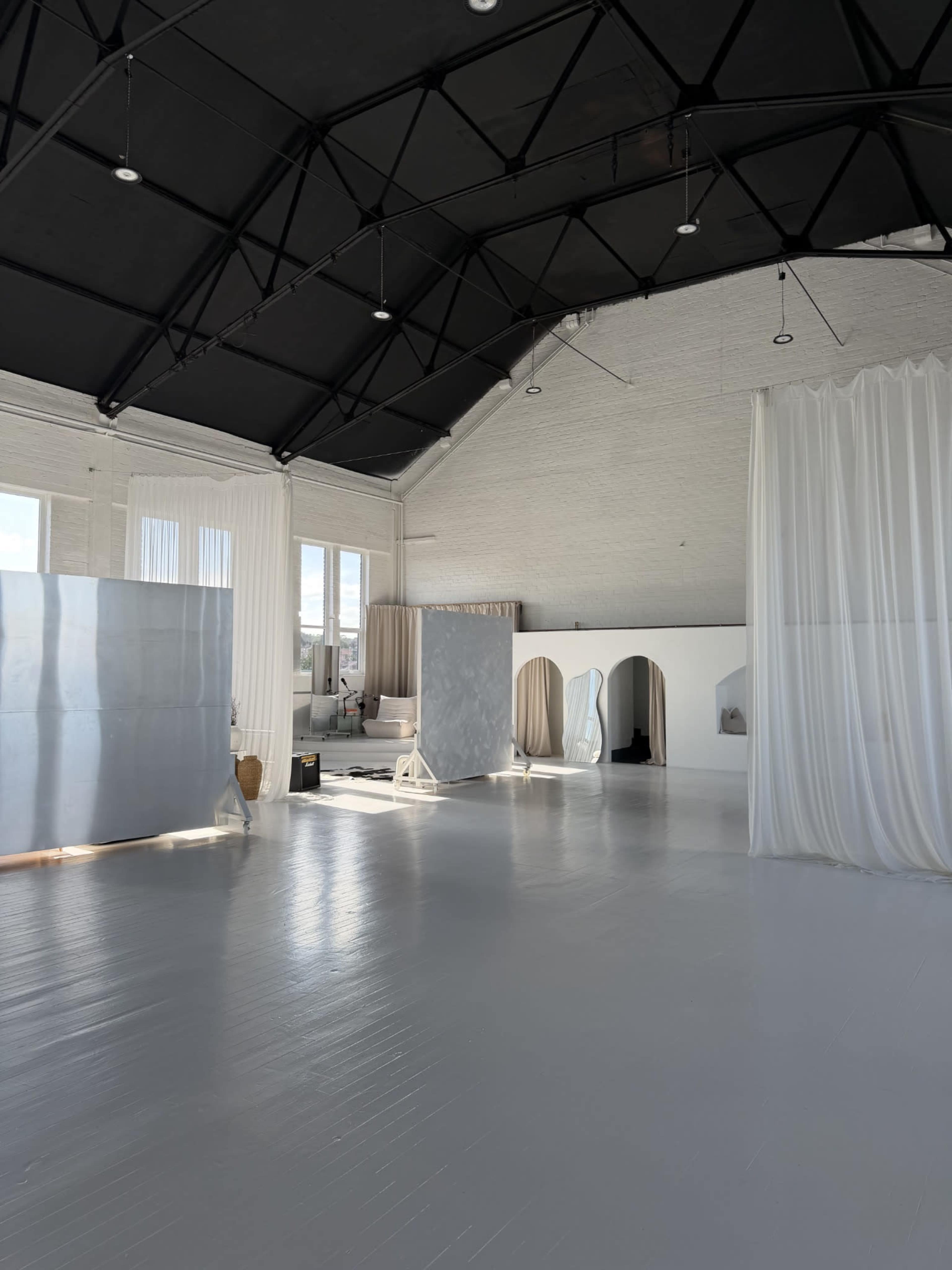 The image shows a spacious, bright interior with a high black ceiling, featuring white walls, sheer curtains, and minimalistic furniture.