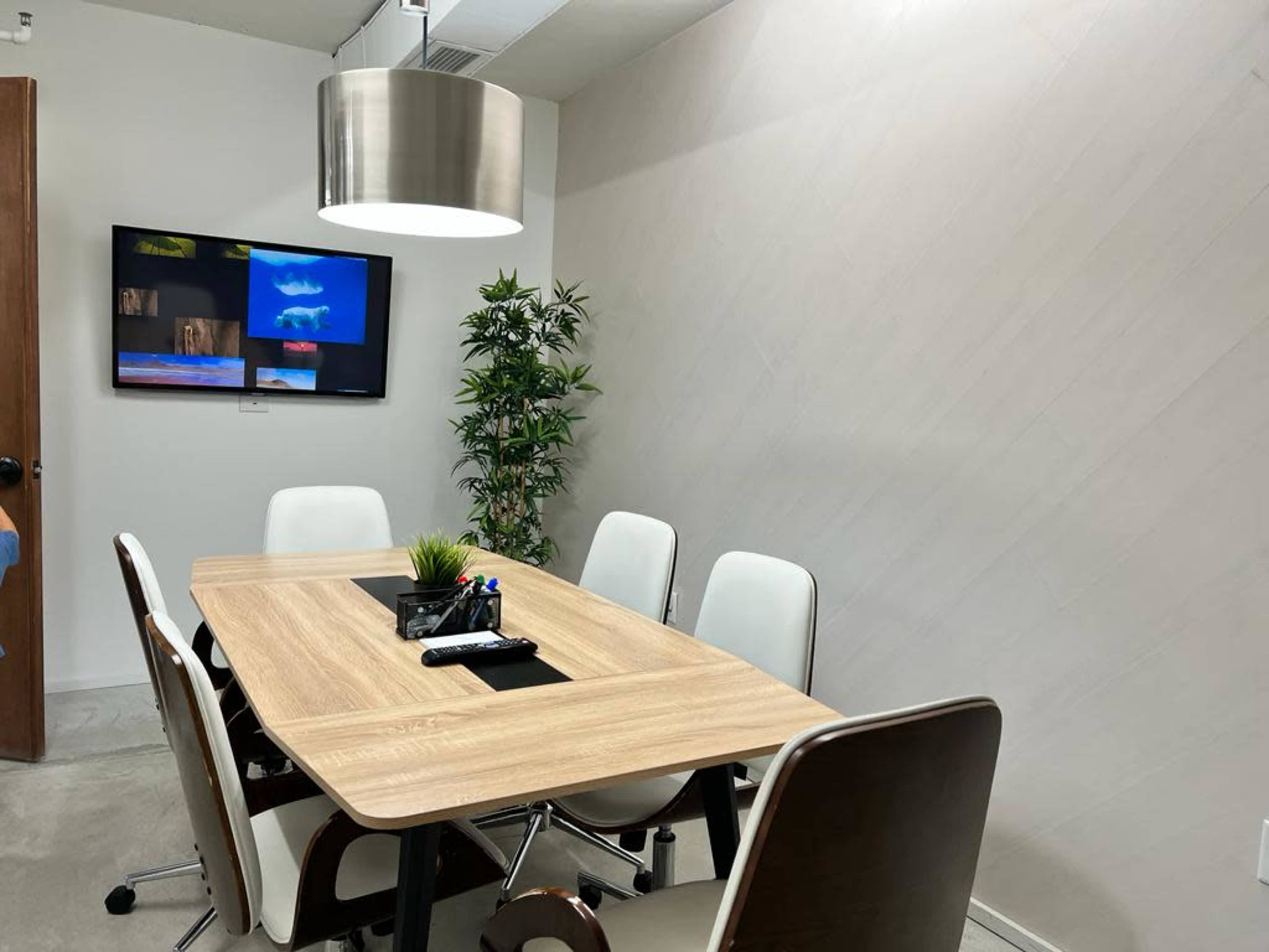 A conference room with a wooden table, white chairs, a plant in the corner, and a wall-mounted screen displaying colorful images.