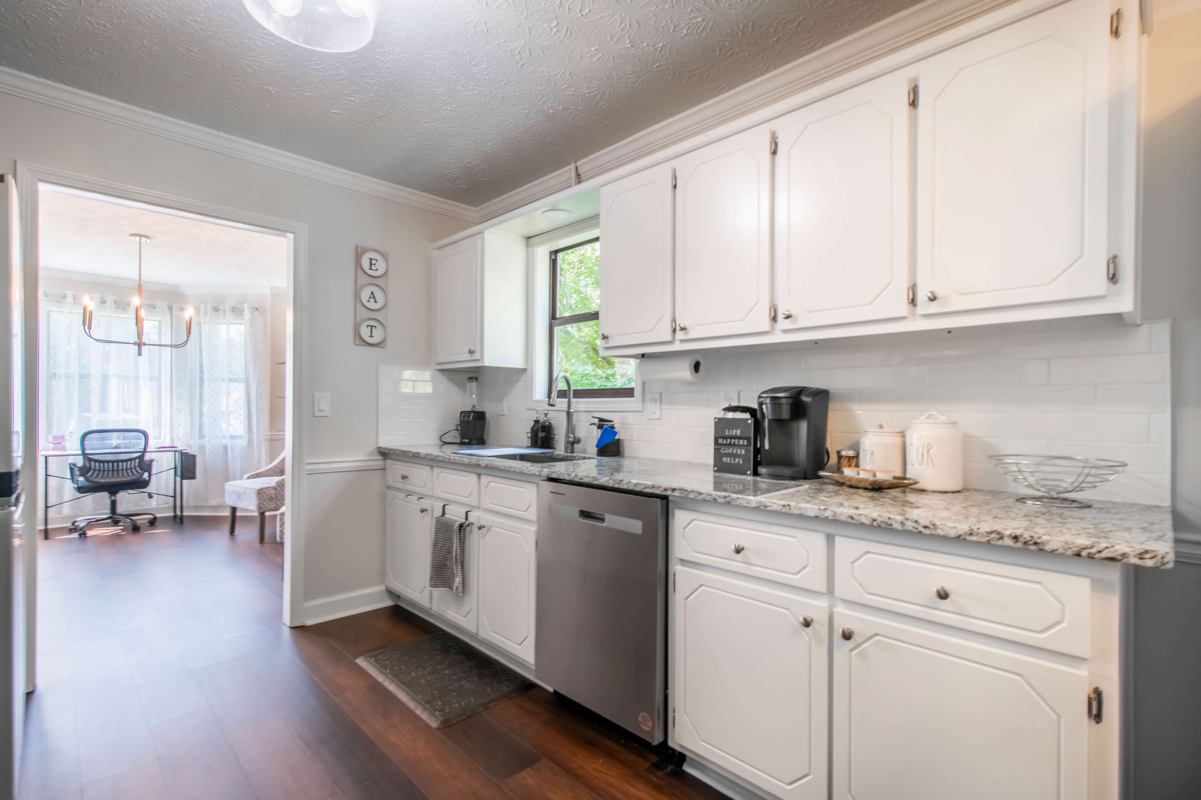 A kitchen with white cabinets, a granite countertop, and a stainless steel dishwasher, leading to a nearby room with a desk and chair.