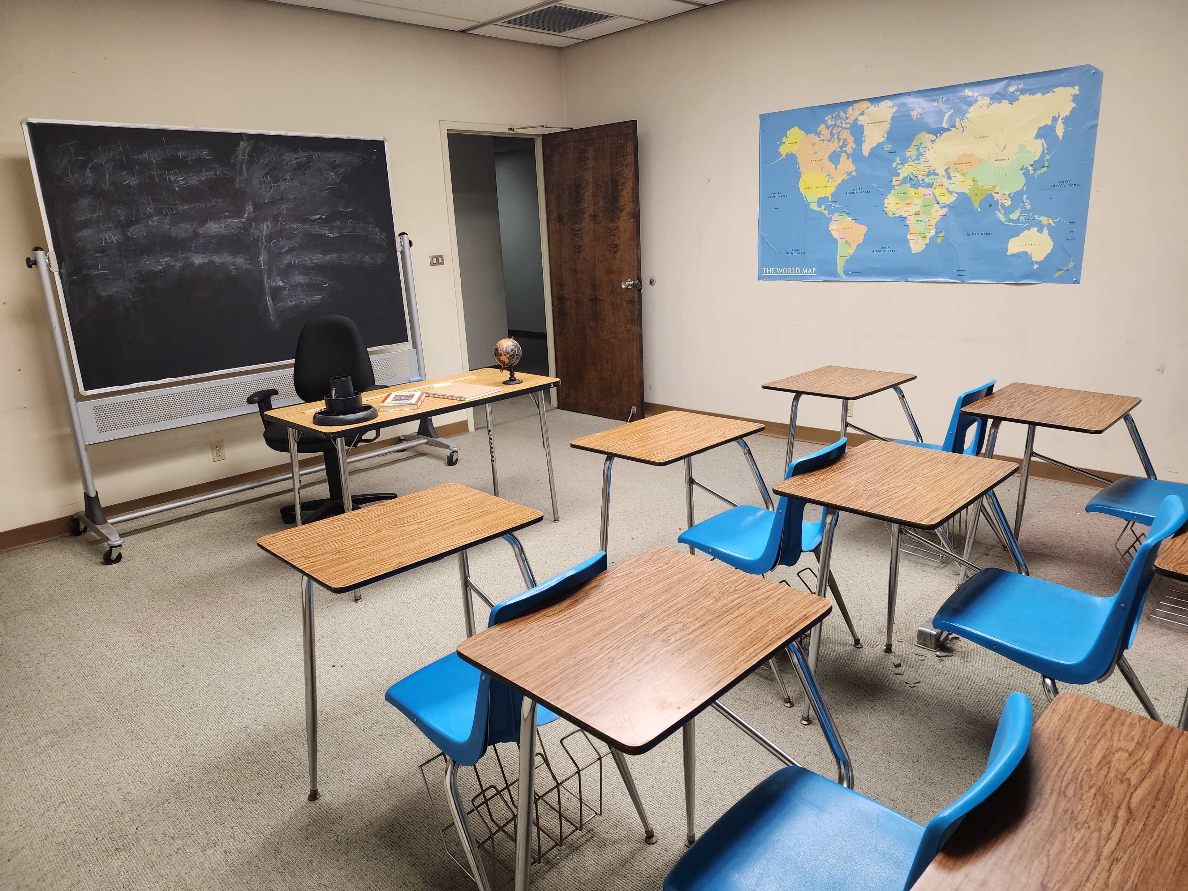 A classroom is furnished with several empty desks, a chair, a globe, and a chalkboard, alongside a world map on the wall.