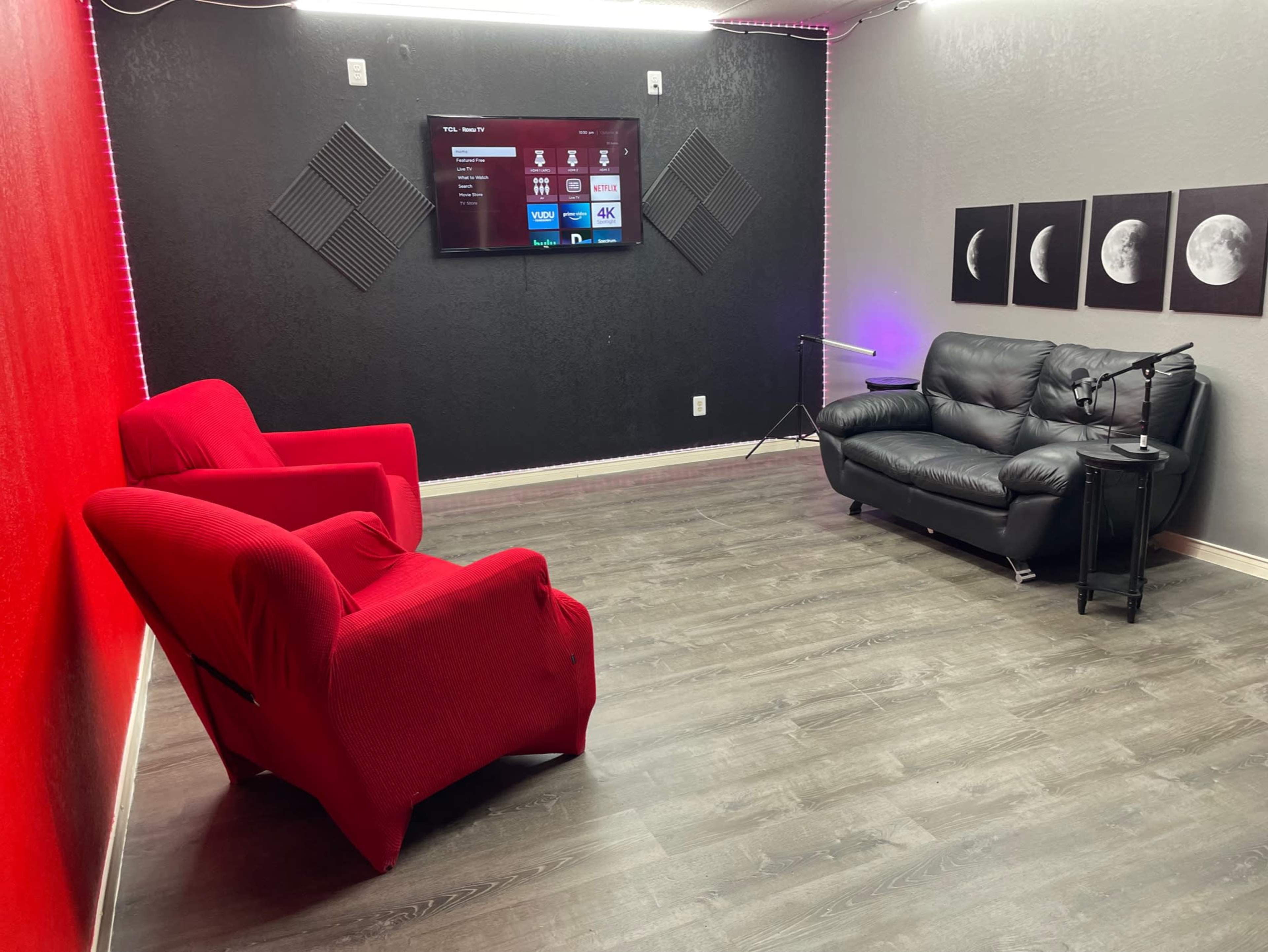 The image shows a small, modern room with a black wall featuring a television, two red chairs, a black sofa, and framed pictures of moons.
