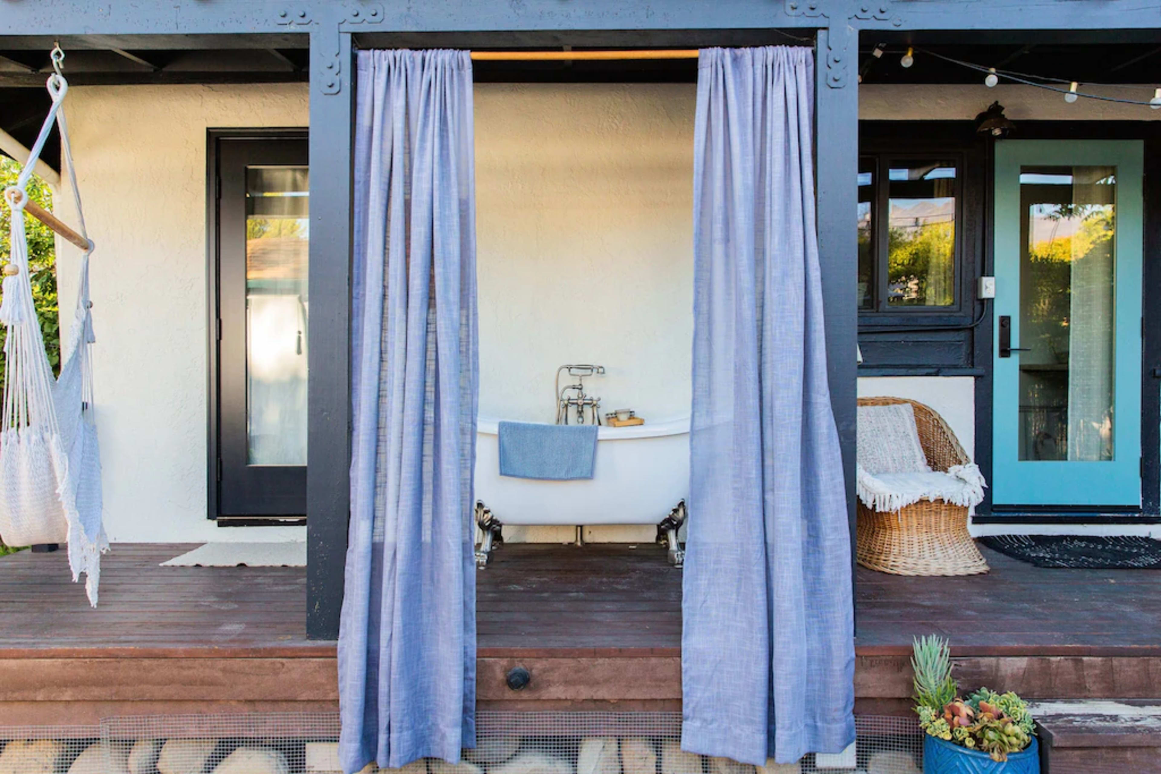 A wooden deck area featuring a vintage bathtub surrounded by draped curtains, with a chair and a plant nearby.