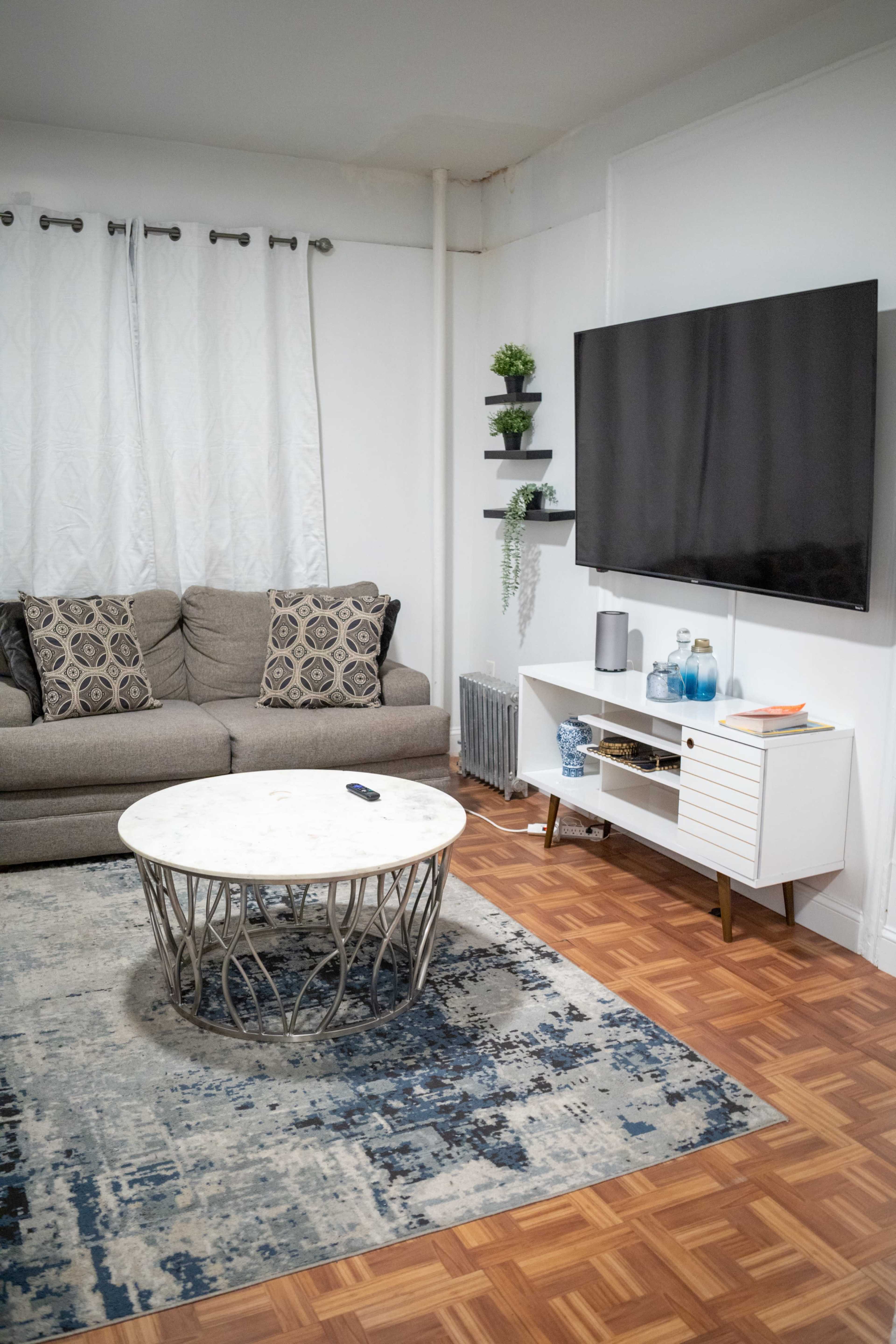 A living room features a gray sofa with decorative pillows, a round marble coffee table, and a television mounted on a white unit, complemented by a blue and beige area rug.