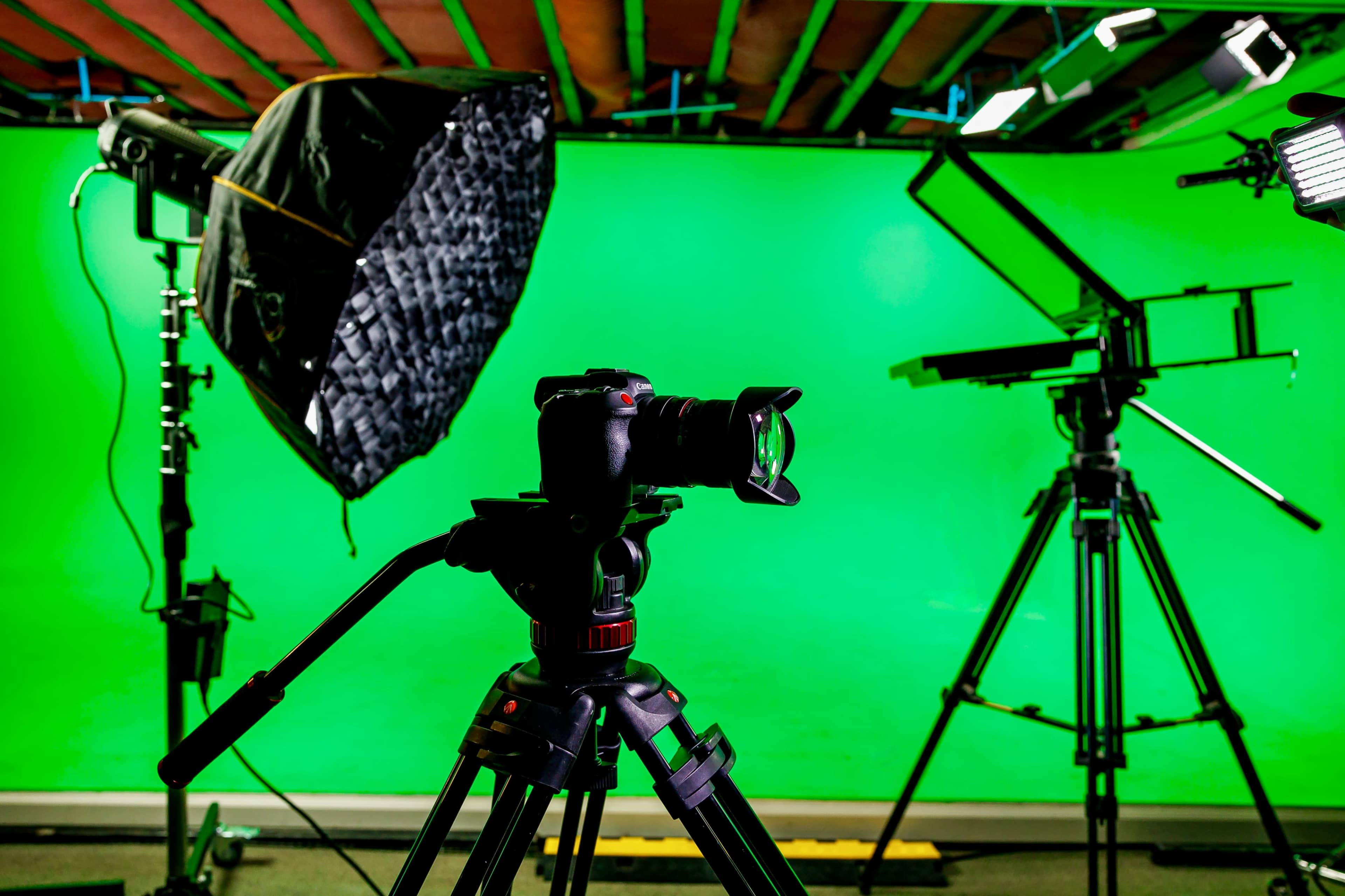 A camera is mounted on a tripod in front of a green screen setup, surrounded by studio lighting equipment.