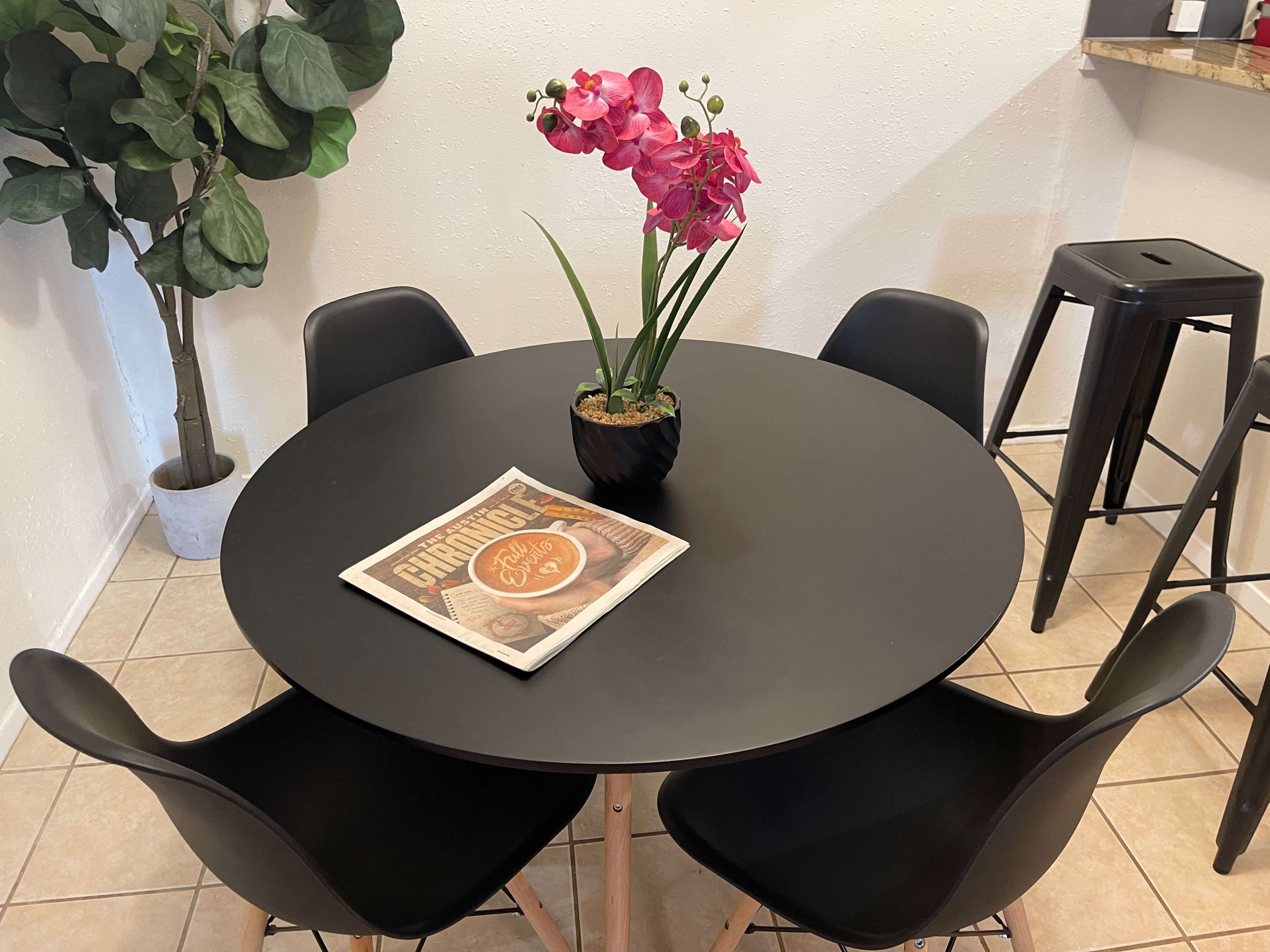 A black round table is set with a potted orchid and a magazine, surrounded by four black chairs, with a tall plant visible in the background.