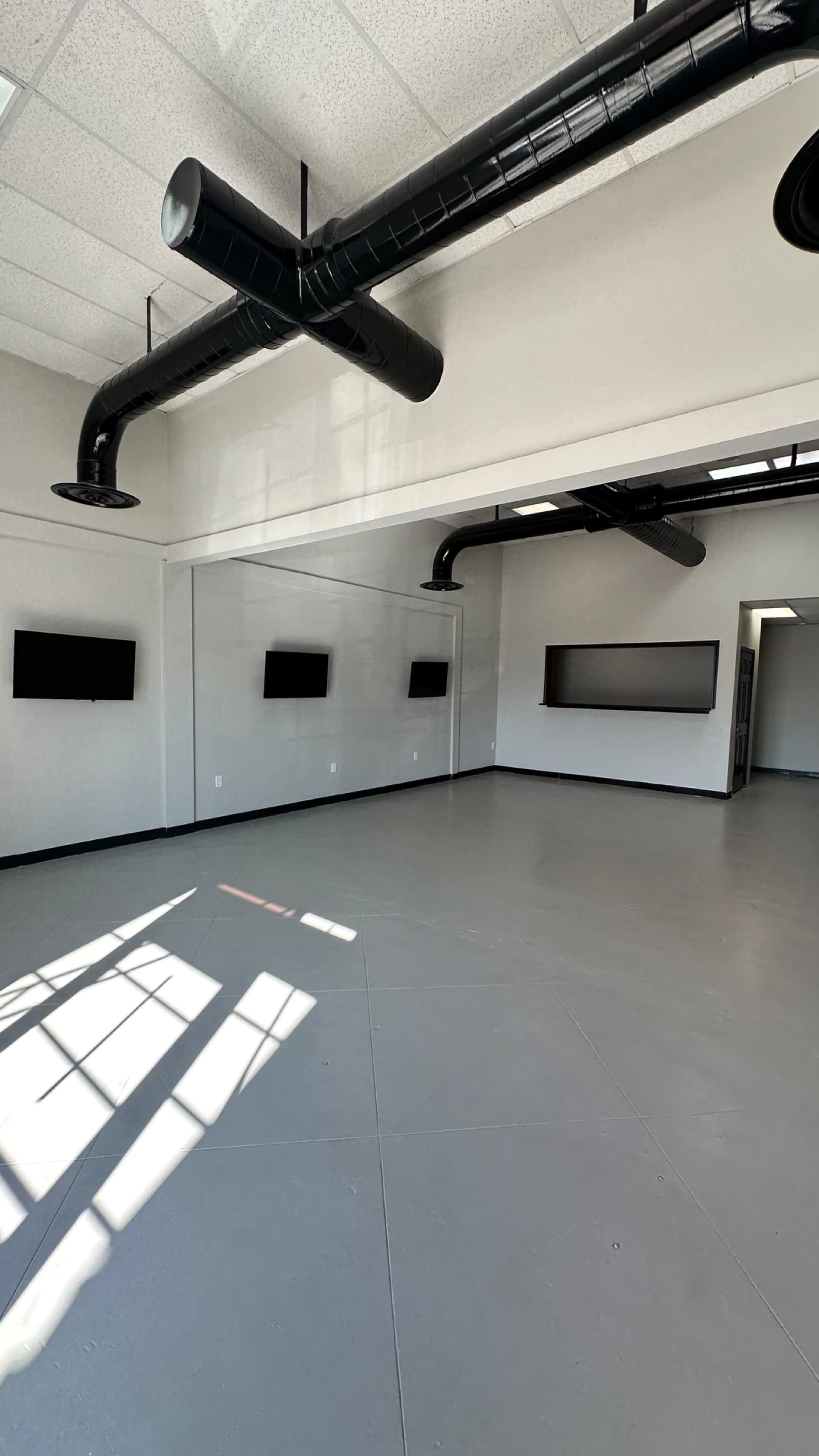 The image shows a spacious, undecorated room with high ceilings, large windows, and exposed black ductwork, featuring several flat-screen monitors mounted on the walls.