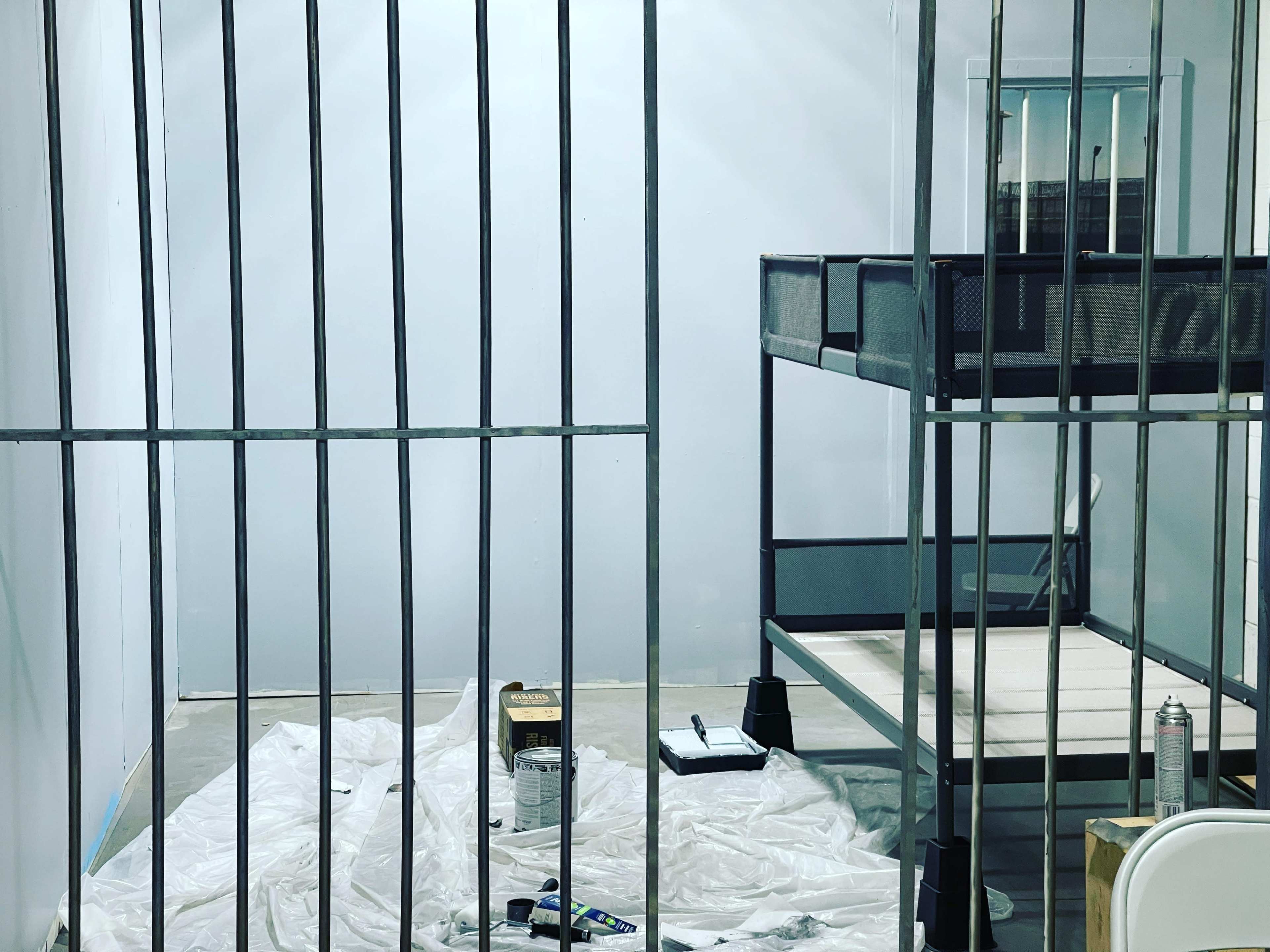 The image shows a partially furnished room with metal bars, featuring a bunk bed against one wall and paint supplies scattered on a tarp-covered floor.