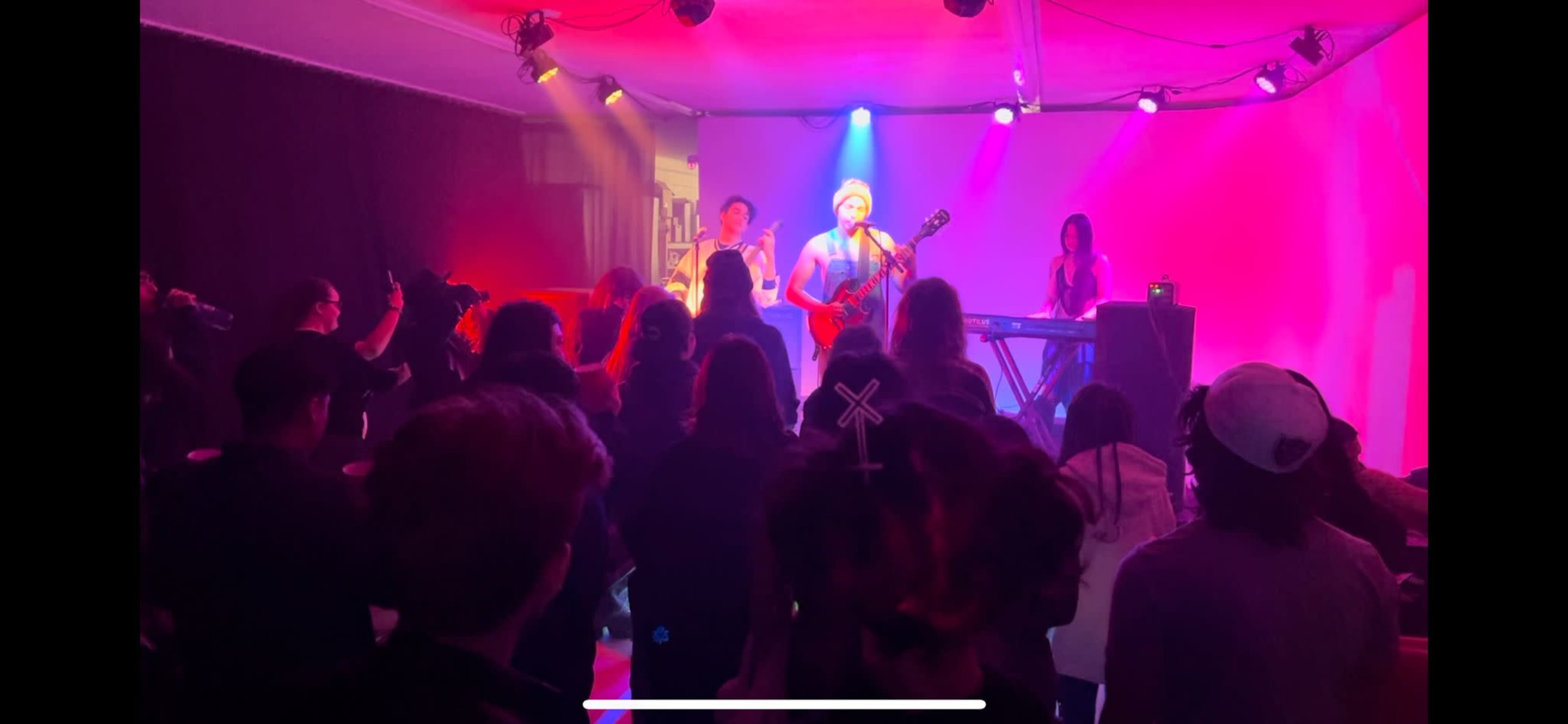 A band performs on stage in front of an audience in a dimly lit venue with colorful lighting.