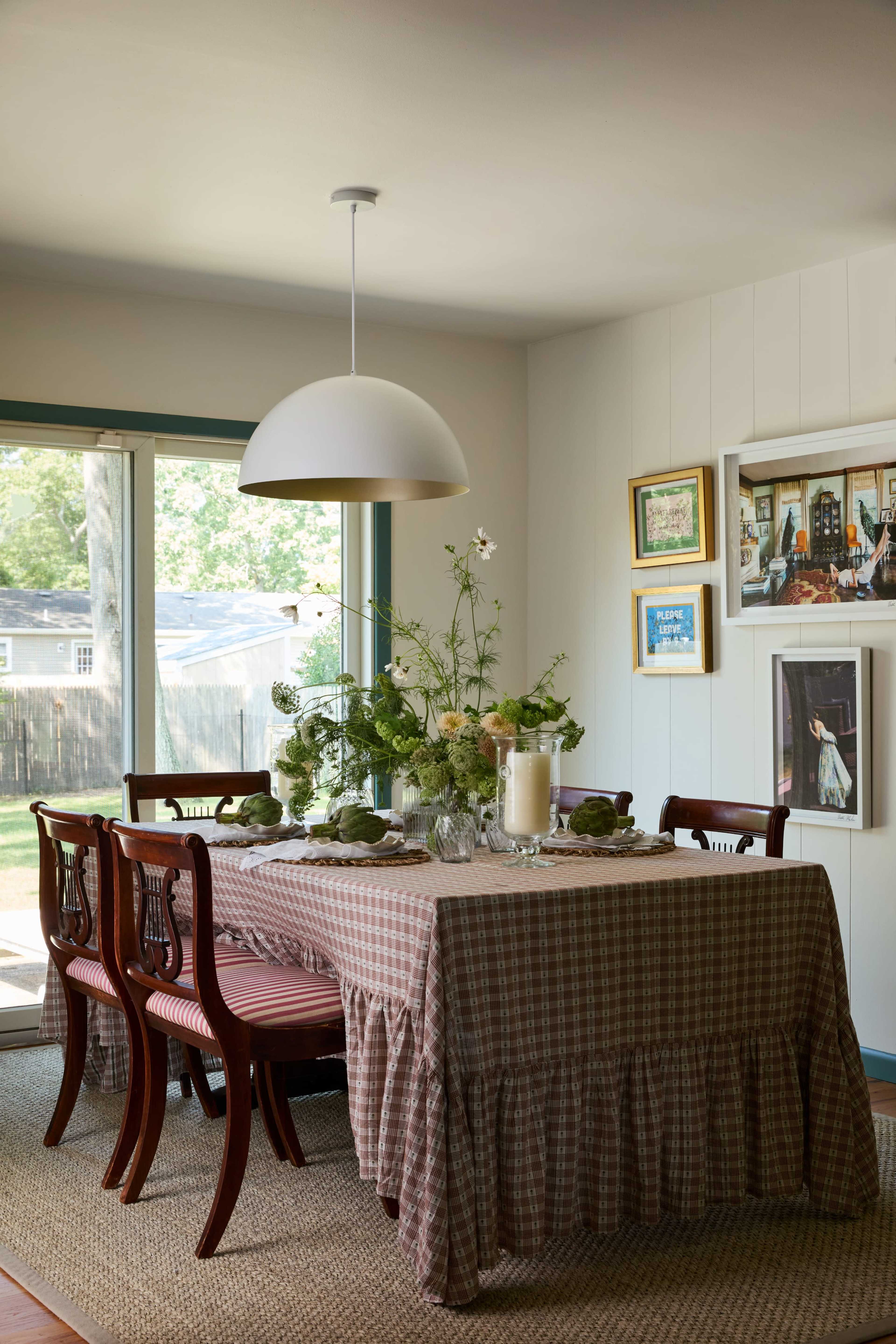 The dining room features a table set with a checked tablecloth, surrounded by wooden chairs, and decorated with flowers and a large pendant light above.