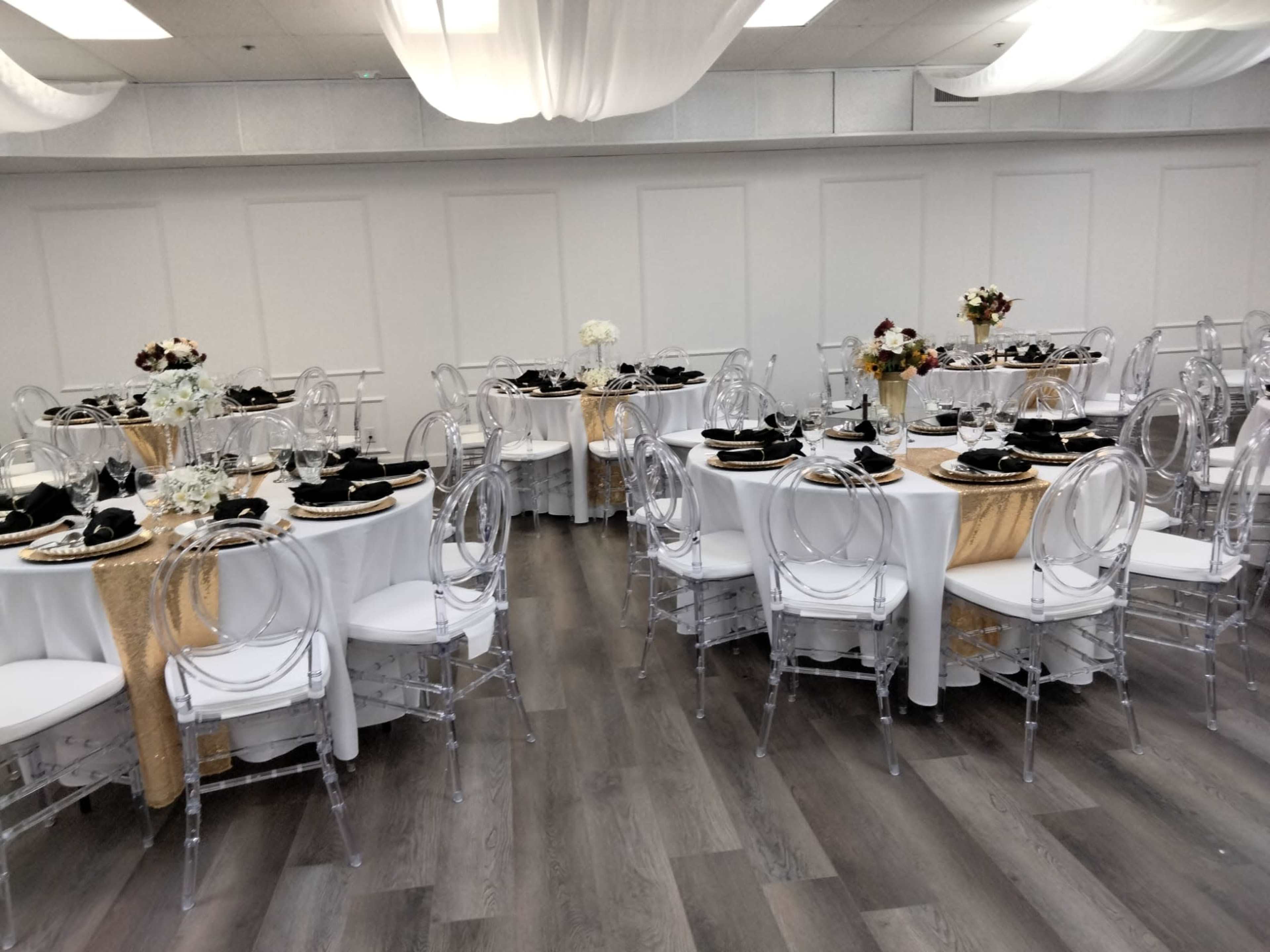 The image depicts a banquet hall set with multiple round tables covered in white tablecloths, adorned with floral centerpieces and black dinnerware, surrounded by transparent chairs.