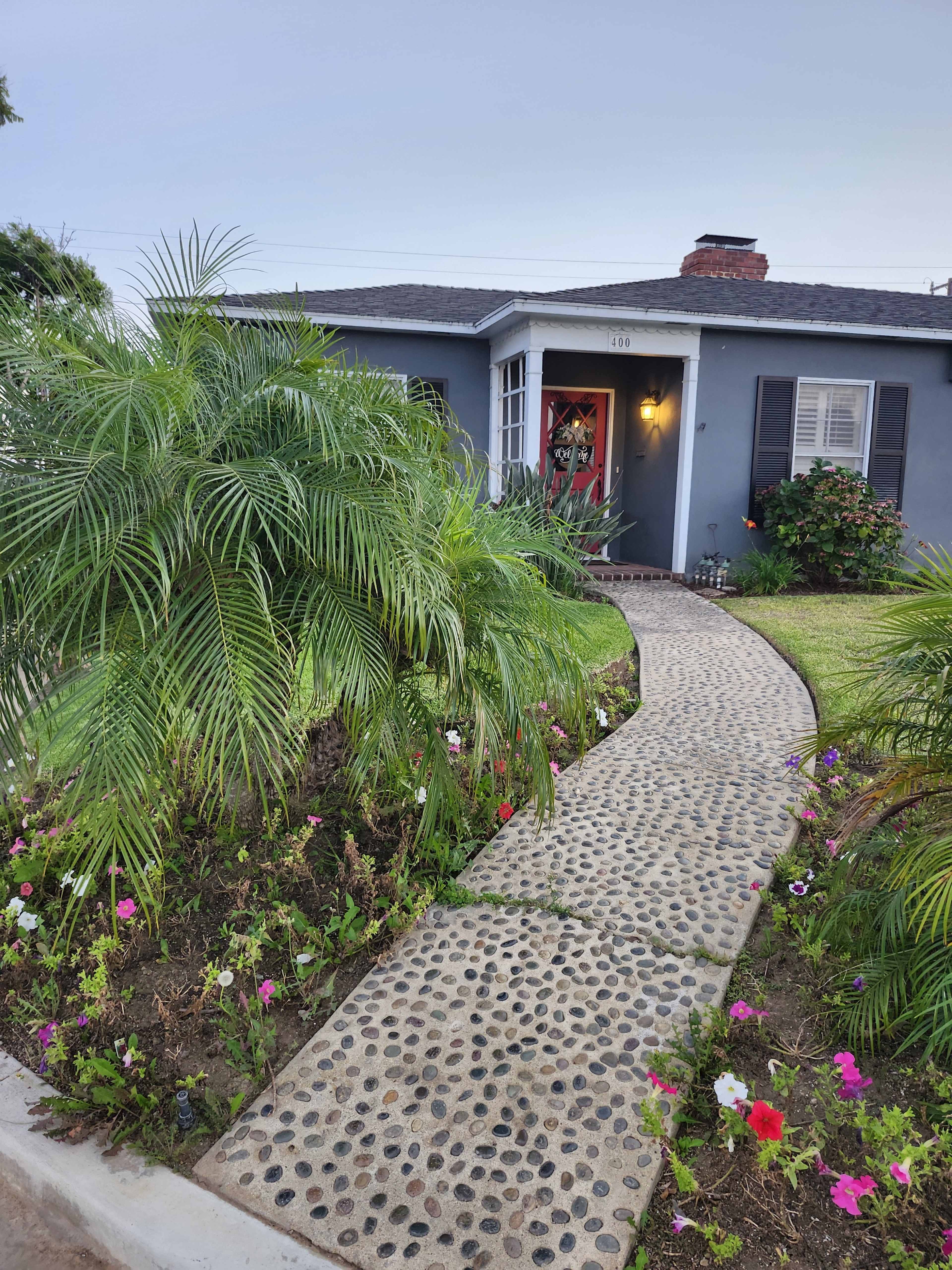 A stone pathway leads to a gray house surrounded by green palm plants and colorful flowers.