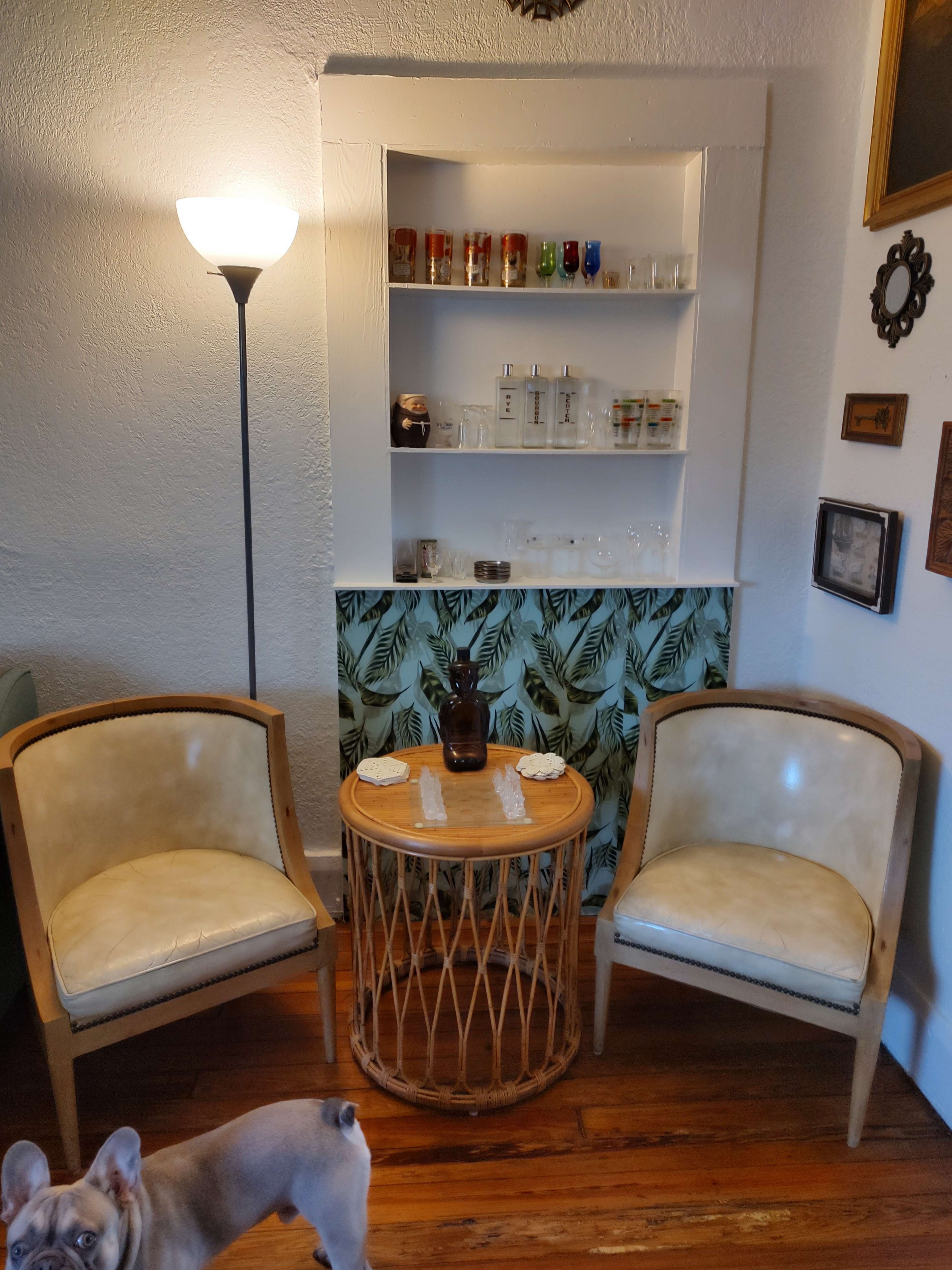 Two circular chairs beside a small round table, with a lamp and a shelf displaying various glass containers in a room decorated with a tropical leaf motif.