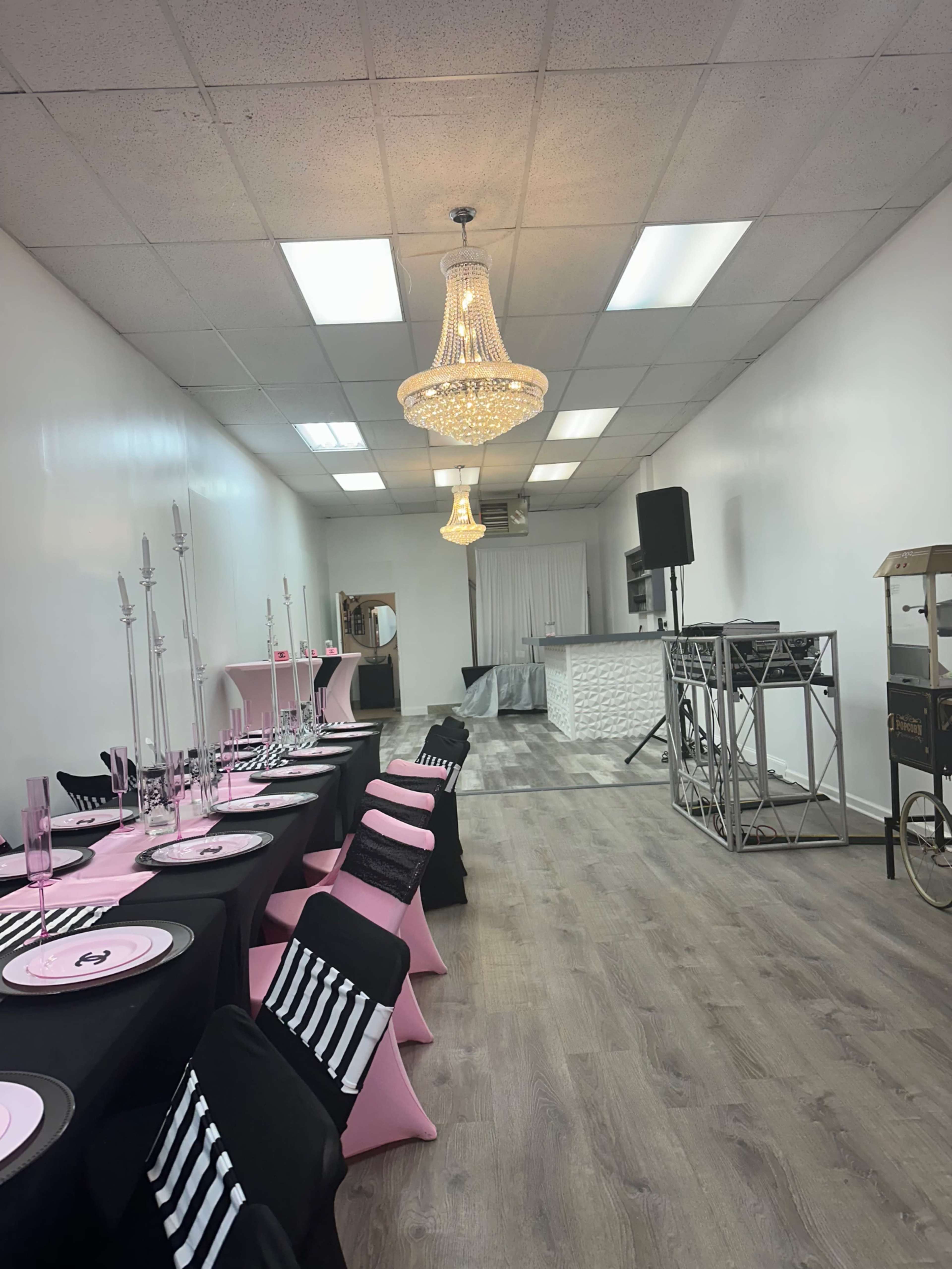 The image shows a bright event space featuring tables with black and pink tablecloths, a stage area at the back, and two chandeliers hanging from the ceiling.