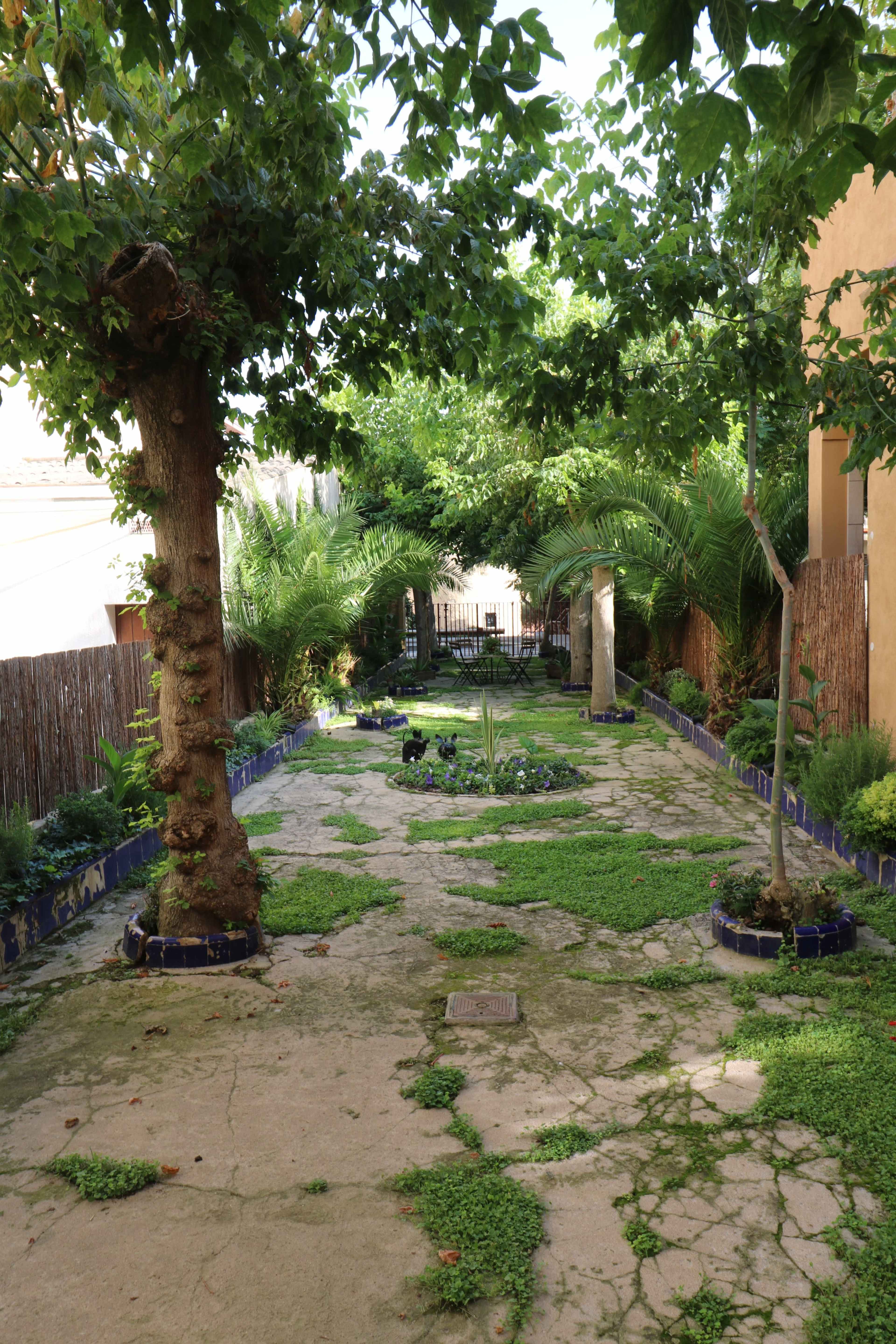 A garden path lined with greenery and stone tiles, leading to a gated entrance framed by trees.